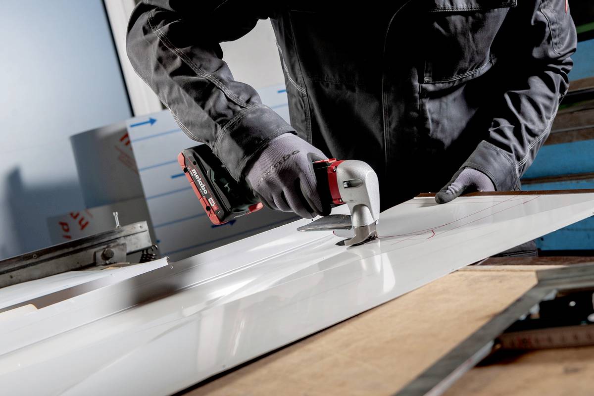 A person is cutting sheet metal with an electric power tool. The scene shows hands, the tool, and the cut in the metal.