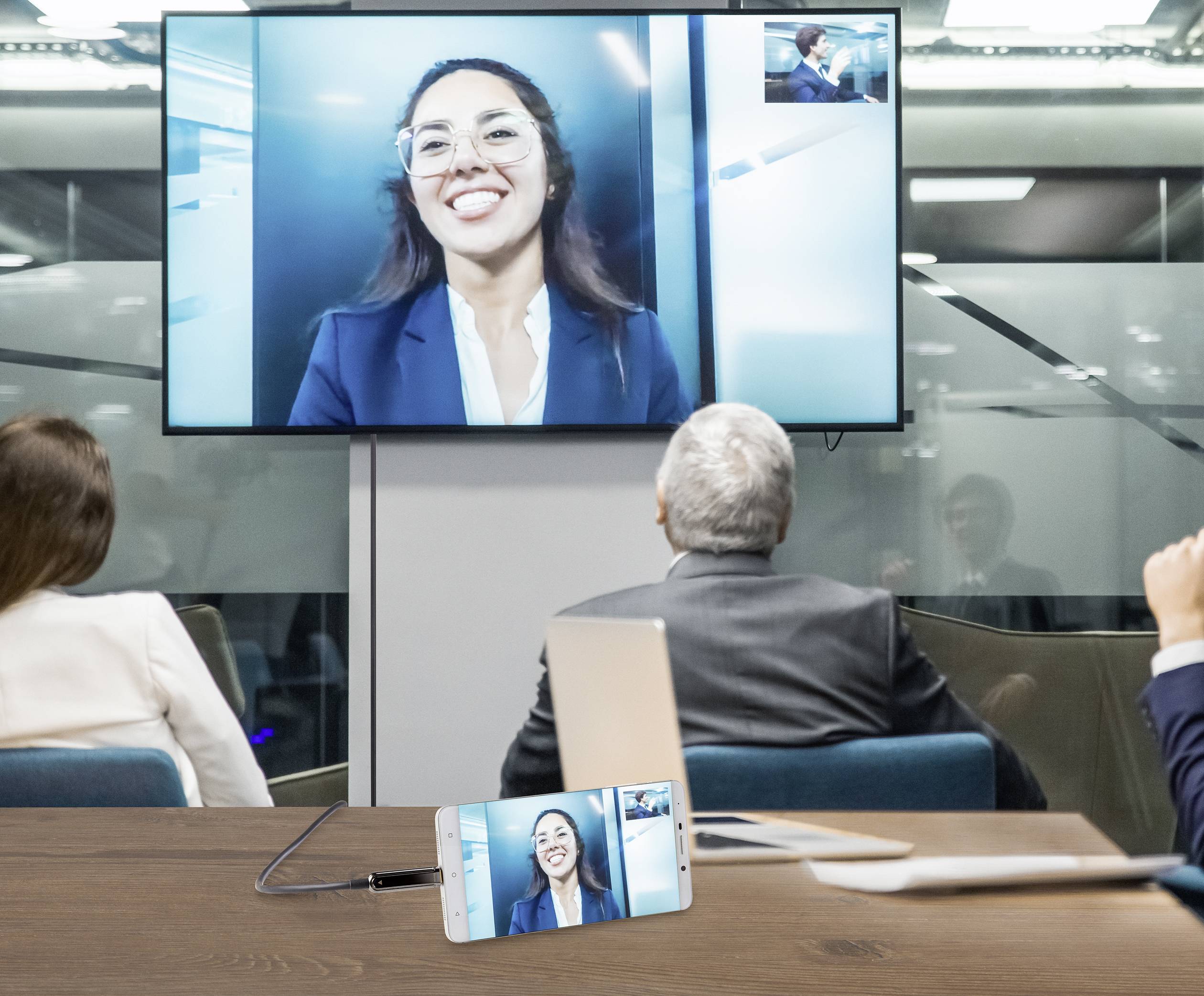 A group of people in a conference room participating in a video conference with a woman on a large screen.