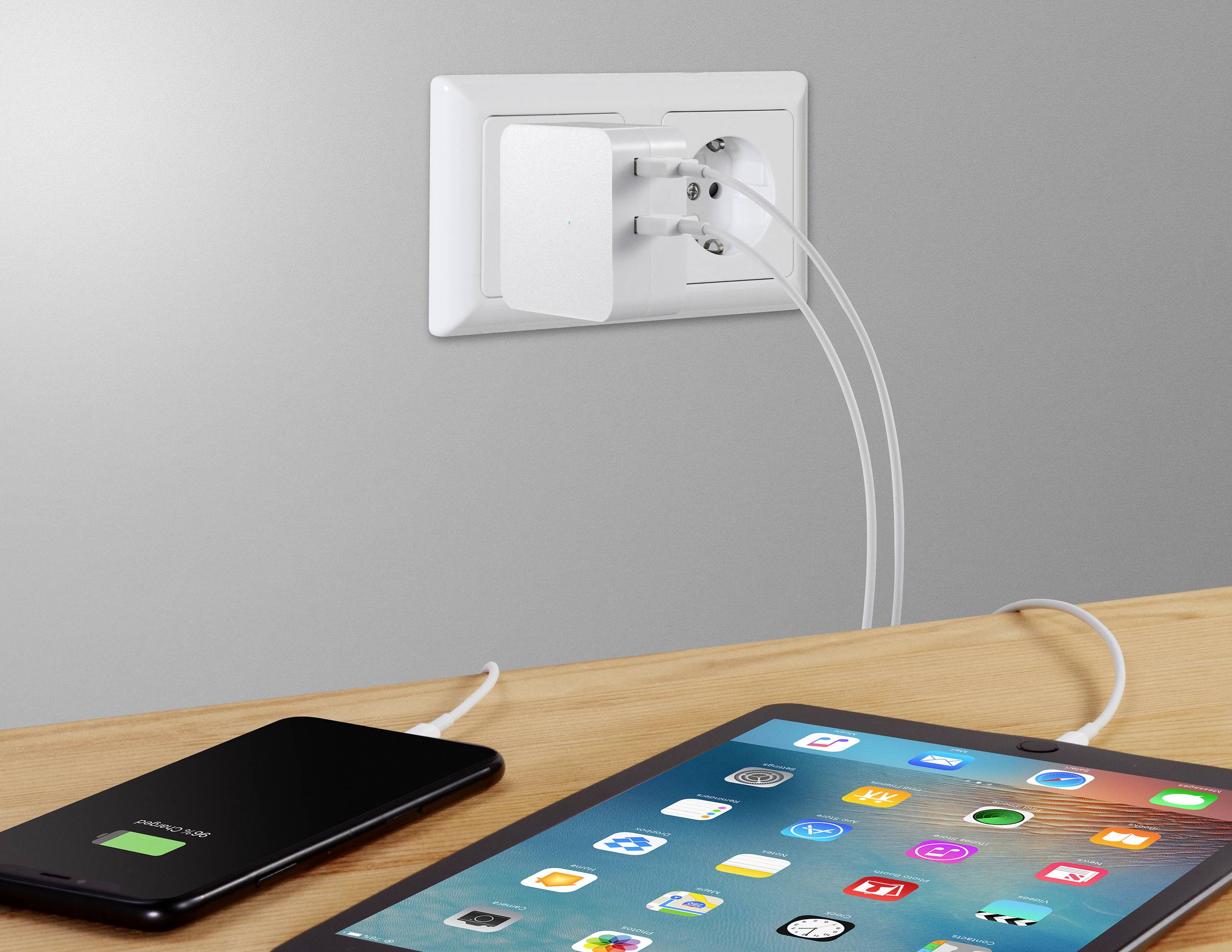 A smartphone and a tablet are being charged on a table, both connected to a double socket in a wall outlet.