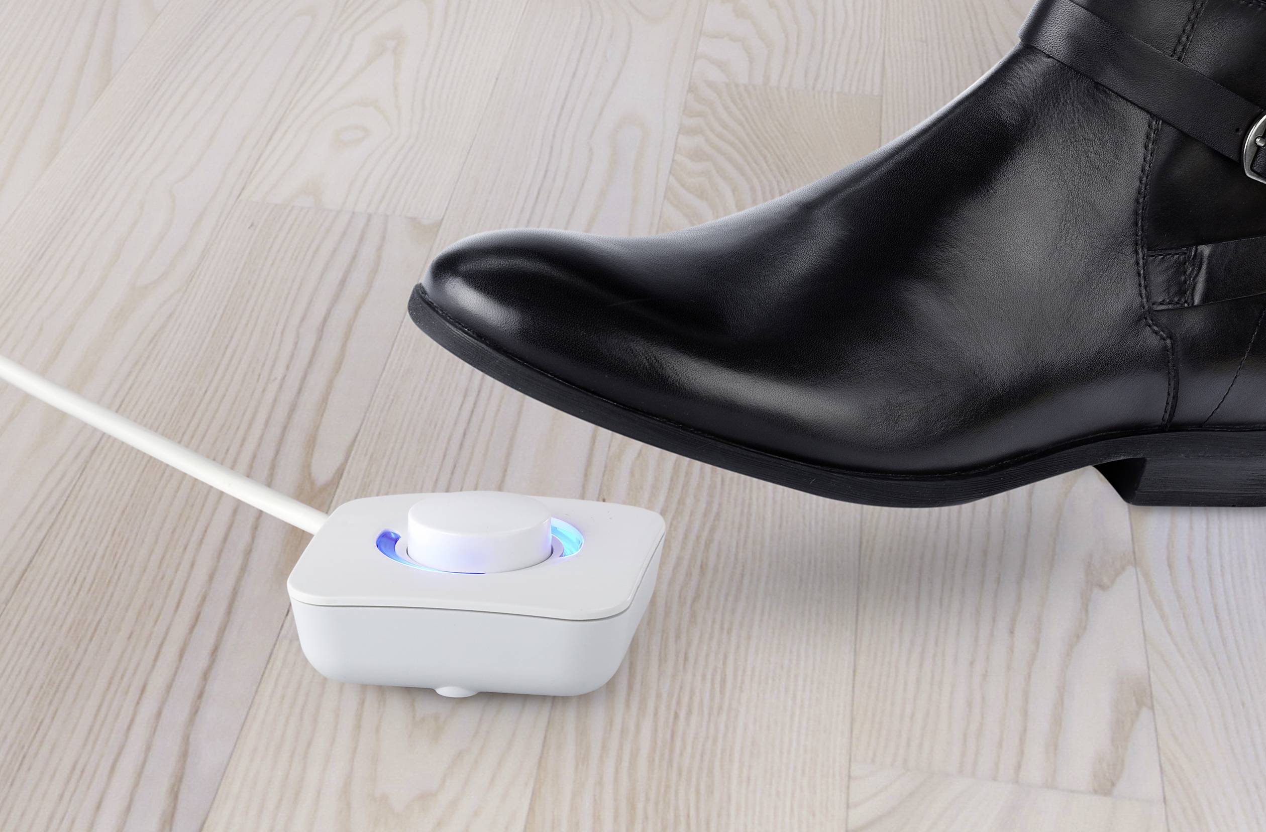 A black shoe steps on a white foot switch with a glowing blue ring on a light wooden floor.