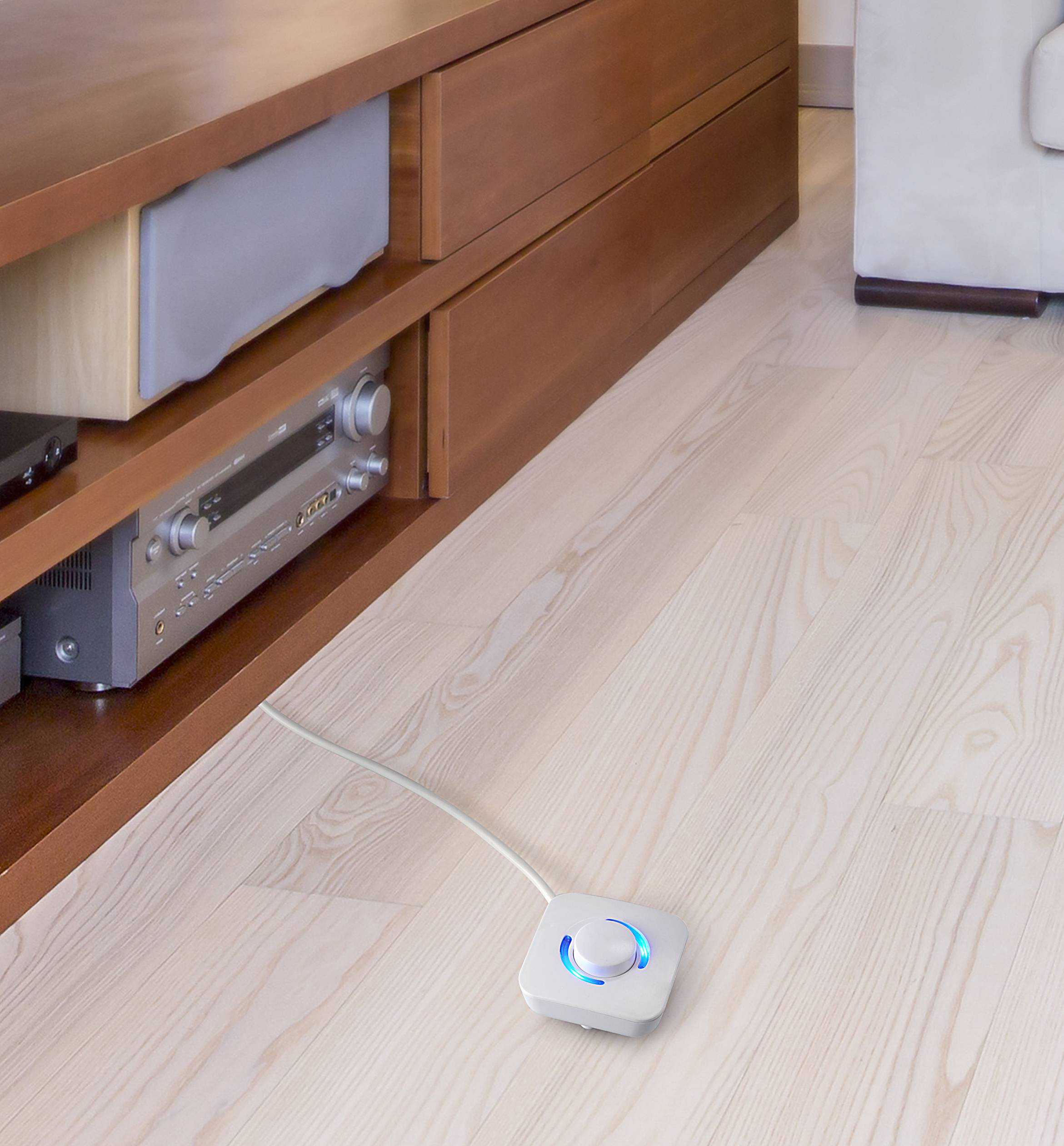 A small, square device with a blue light is resting on a light wooden floor, connected by a cable to a piece of furniture.