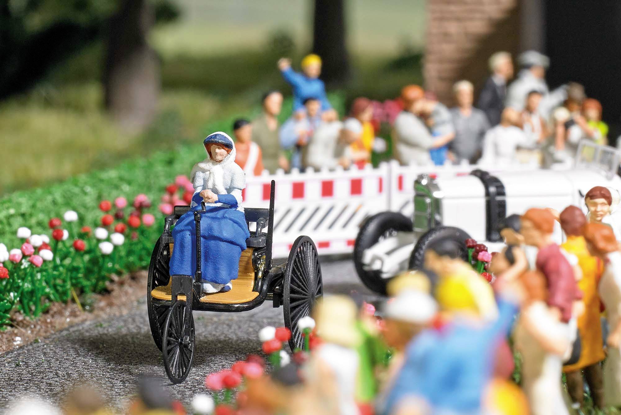 A miniature figurine of a woman in blue clothing driving a three-wheeled vehicle, surrounded by a crowd and colourful flowers.