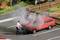 A man is holding a fire extinguisher and standing in front of a red car in a car park, with smoke rising from its engine compartment.