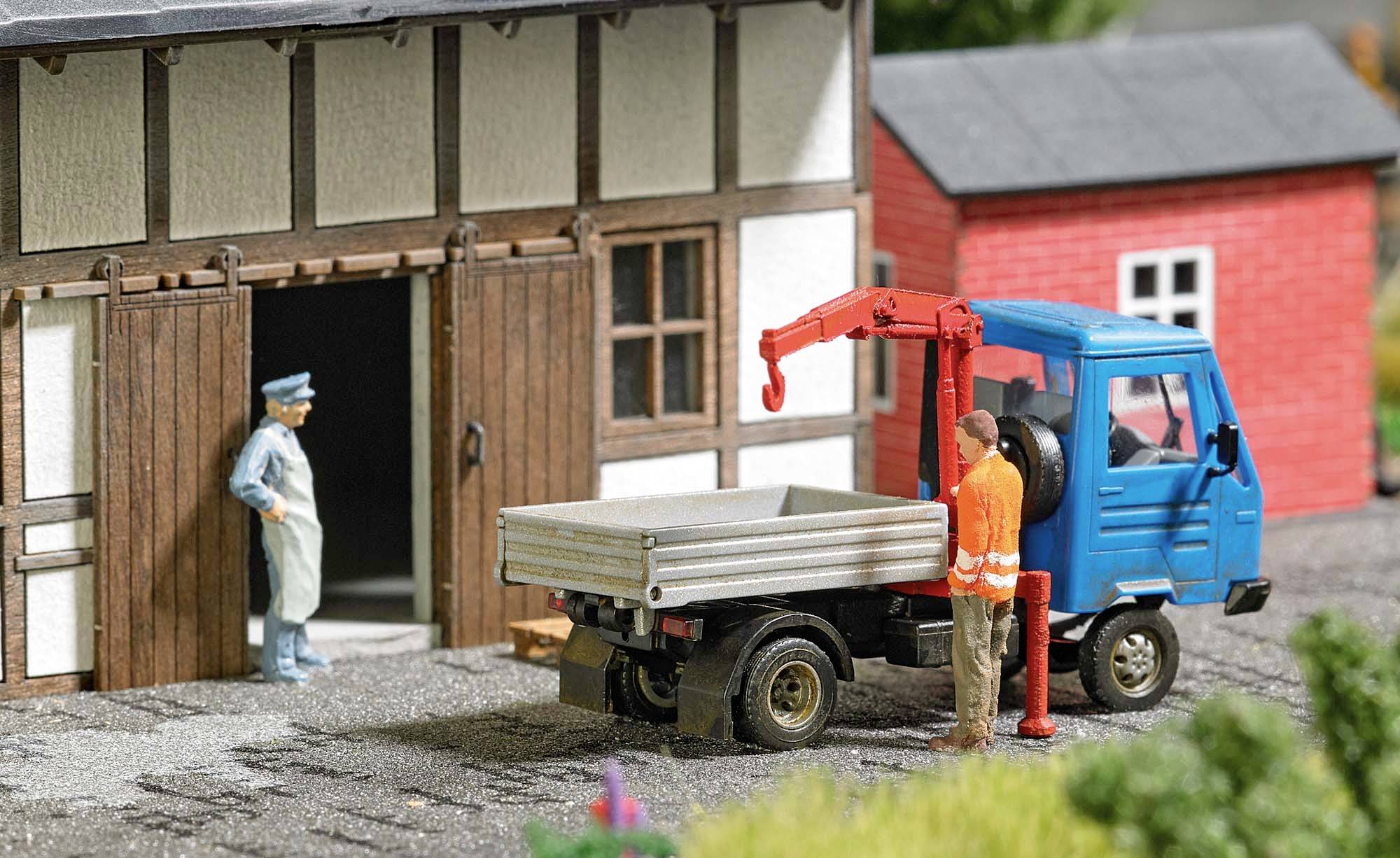 A small blue lorry is parked in front of a barn door. Two men, one in uniform and one in workwear, are having a conversation.