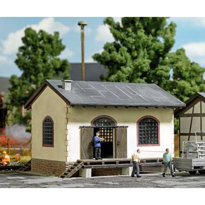 A model of a small warehouse with two figures on the veranda. Trees and construction site activity in the background.
