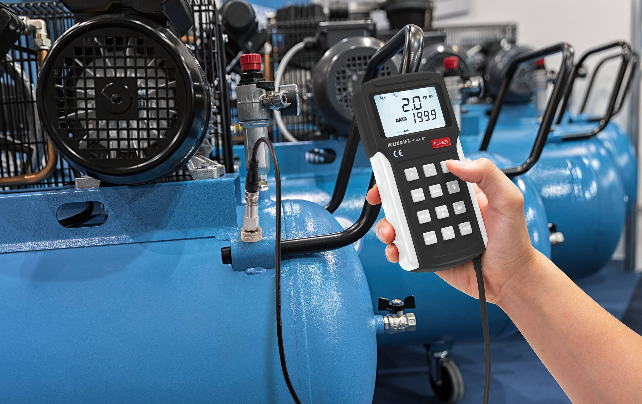A hand is holding a measuring device in front of a blue air compressor. The screen displays '2.0', indicating the pressure.