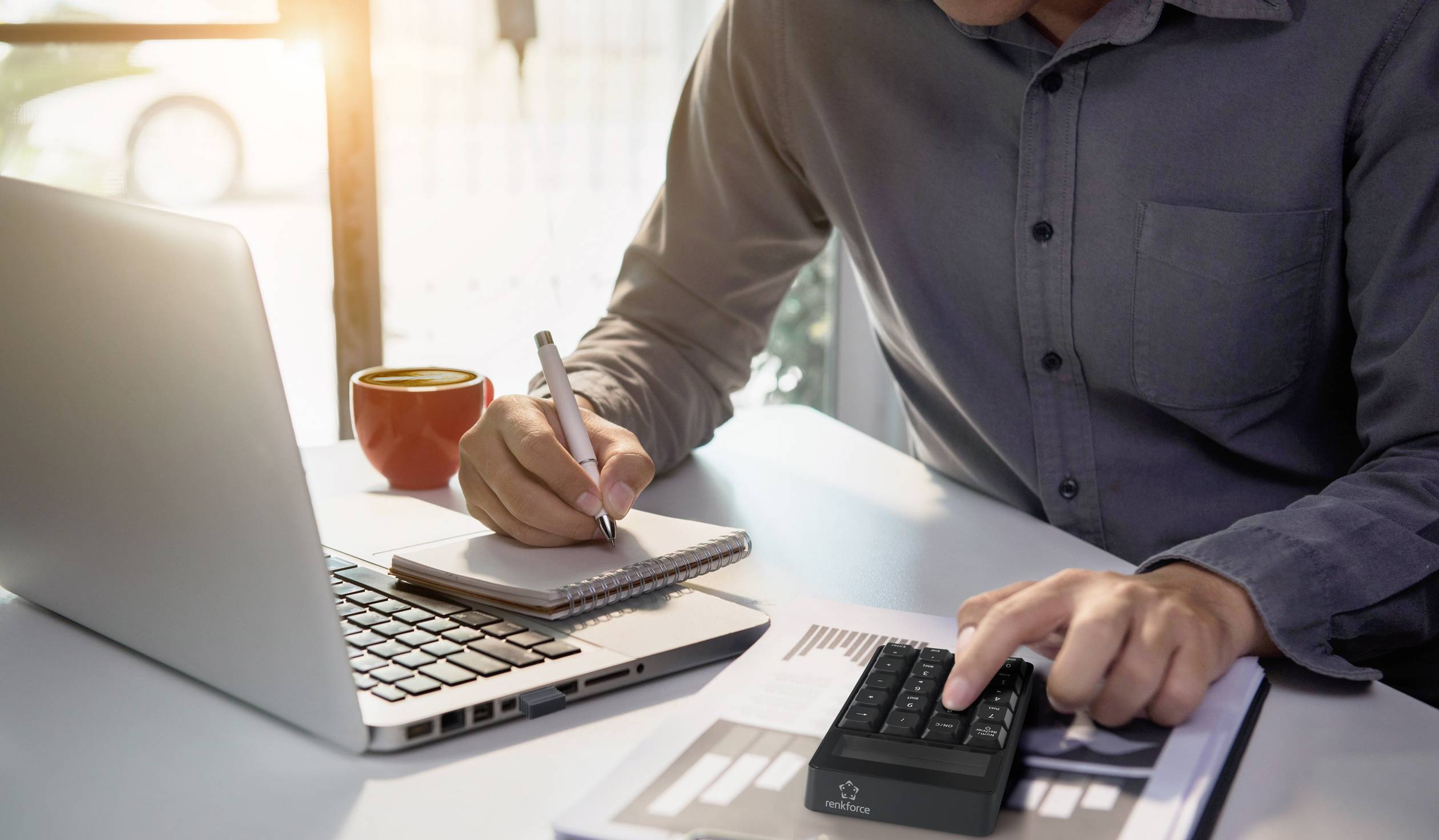 A person is working on a laptop, taking notes on a pad and using an external keyboard. A red mug is sitting next to the laptop.