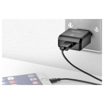 A black USB charger plugged into a wall outlet charges a smartphone on a white table in the foreground.