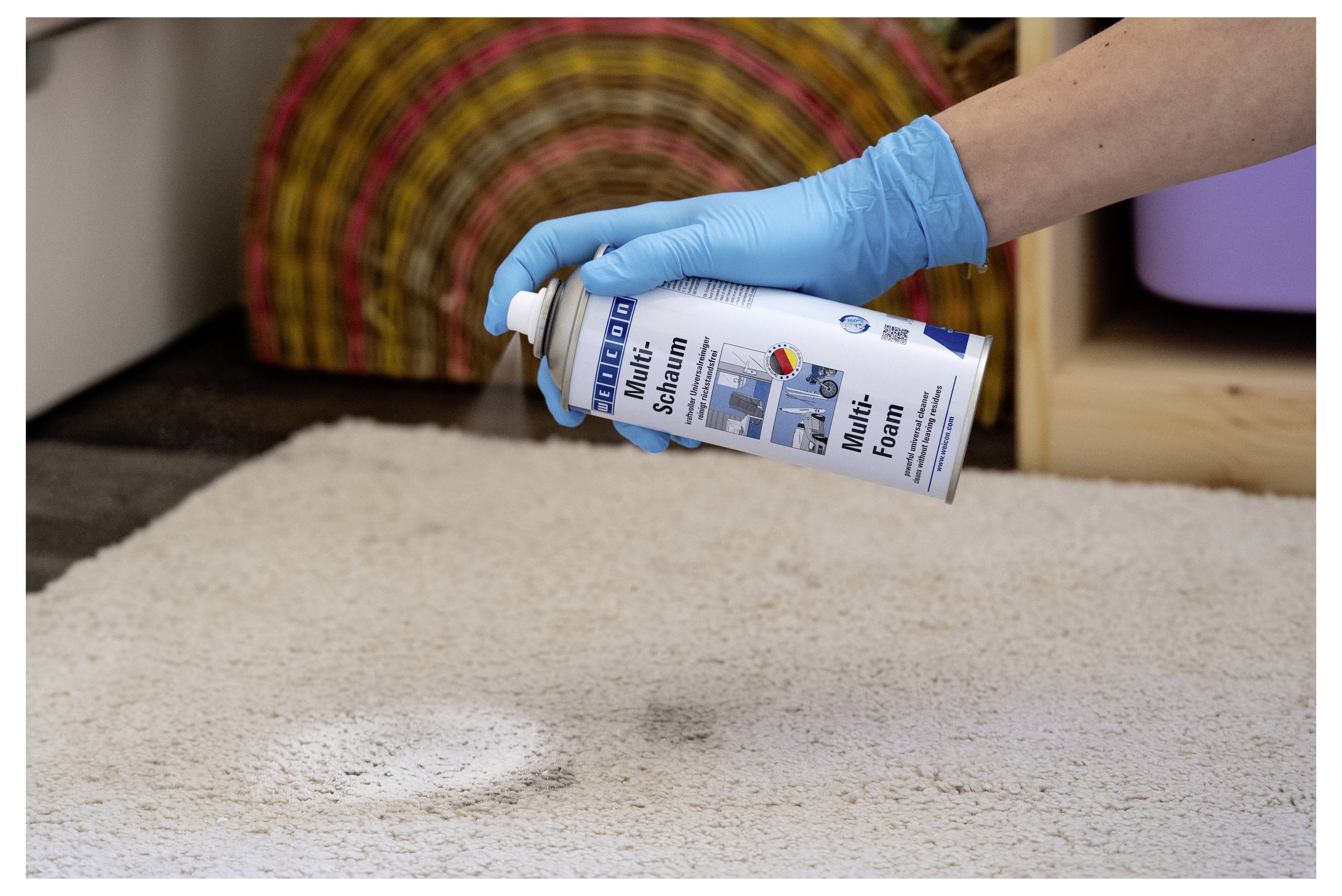 Gloved hand sprays 'Mellerud Multi-Schaum' cleaner onto a dirty white carpet near a rug, treating a stain.