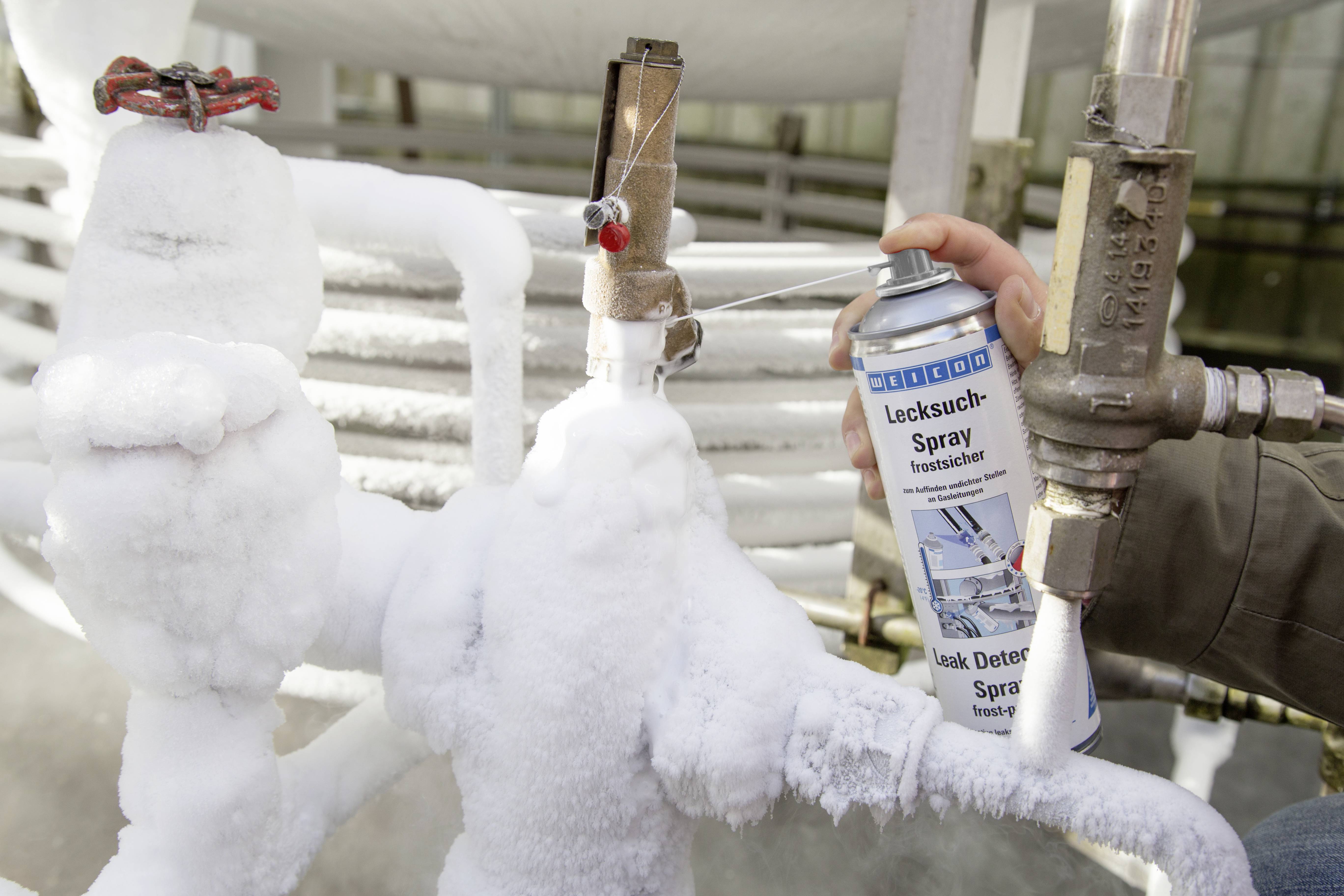A person is spraying a leak detector on frozen pipes to locate leaks. The pipes are heavily covered in ice.