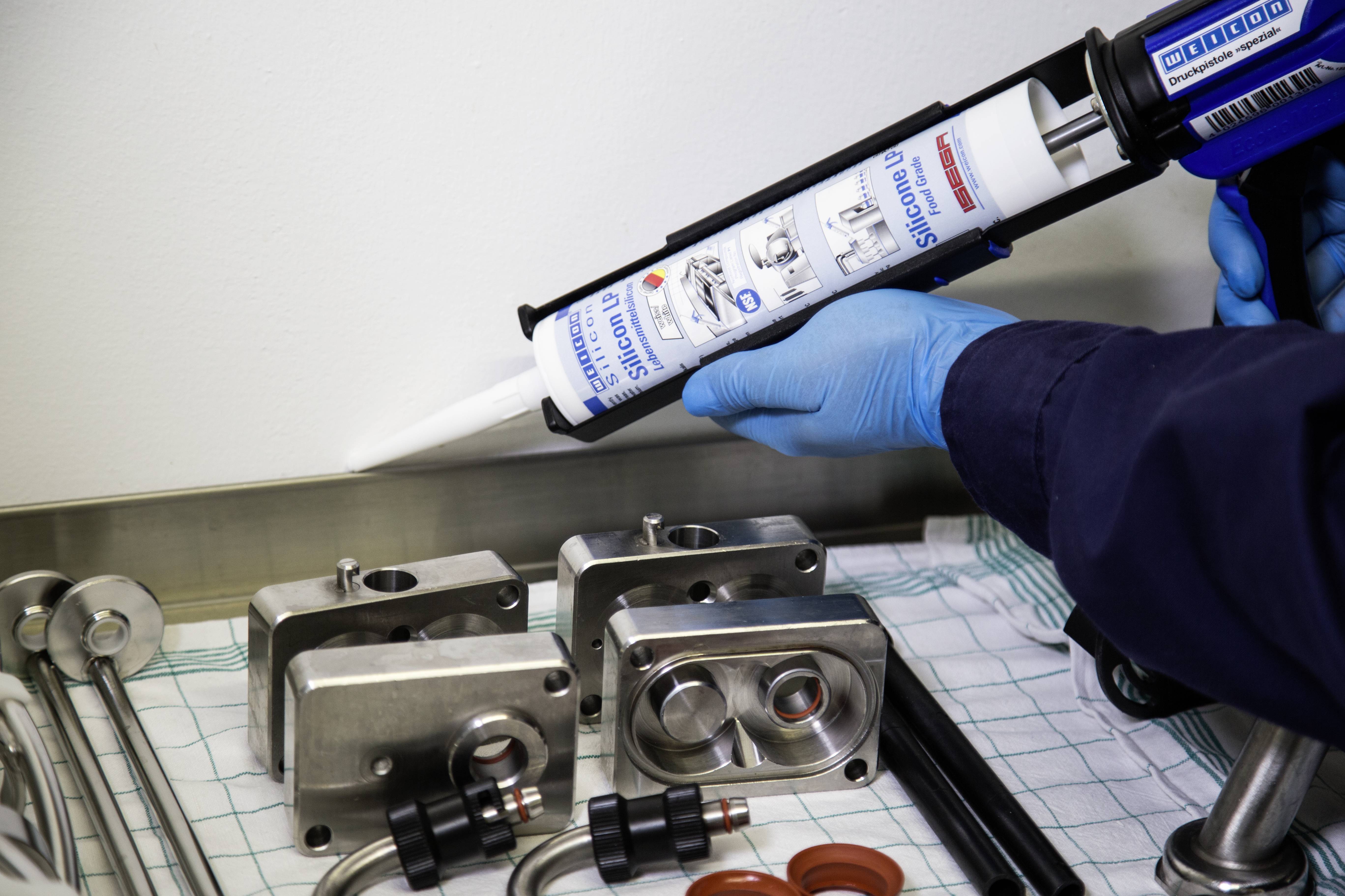 A person is applying sealant using a cartridge gun. Metal parts and tools are lying on the table.