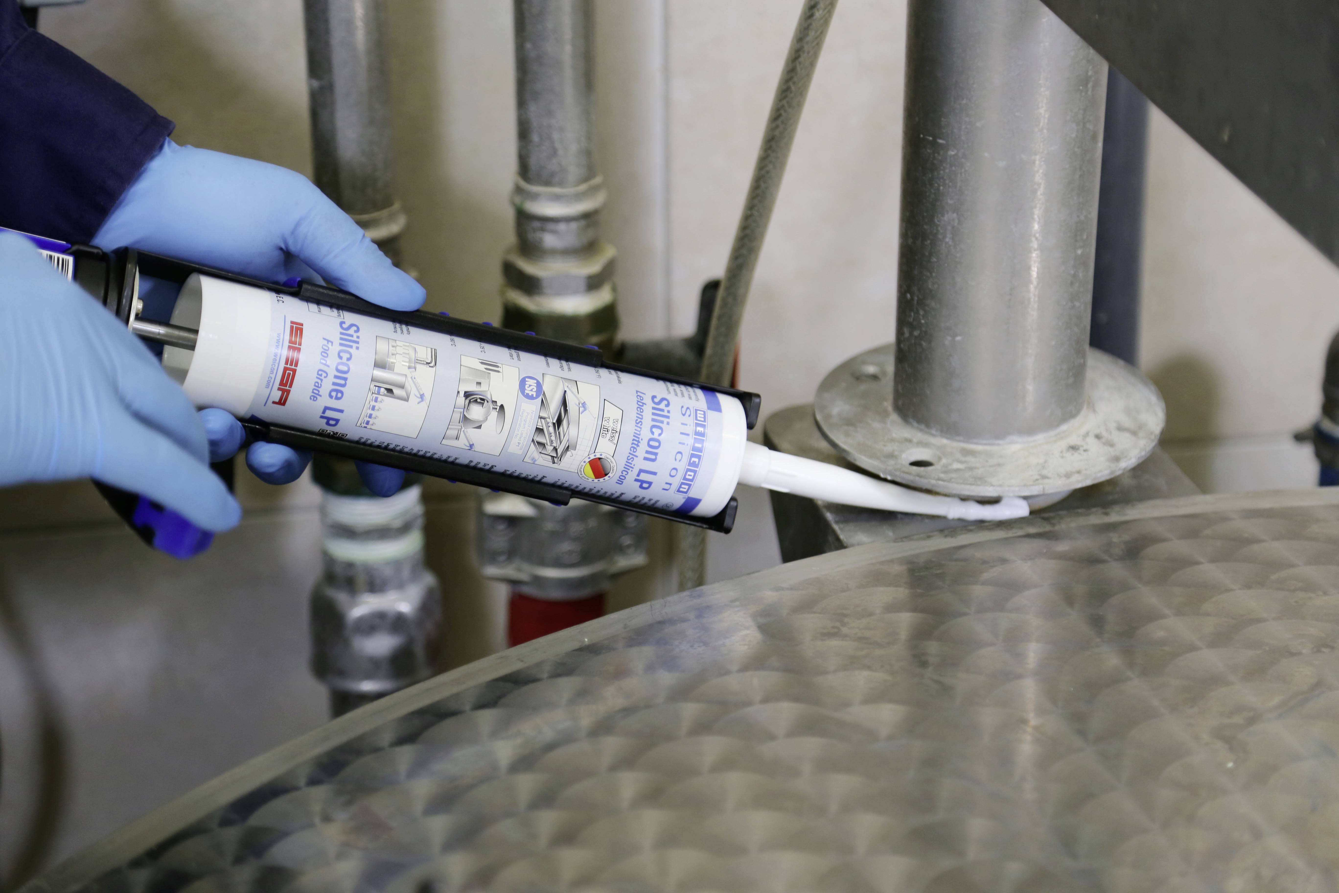 A person is installing a pipe while wearing blue gloves, using sealant from a cartridge to seal a metal connection.