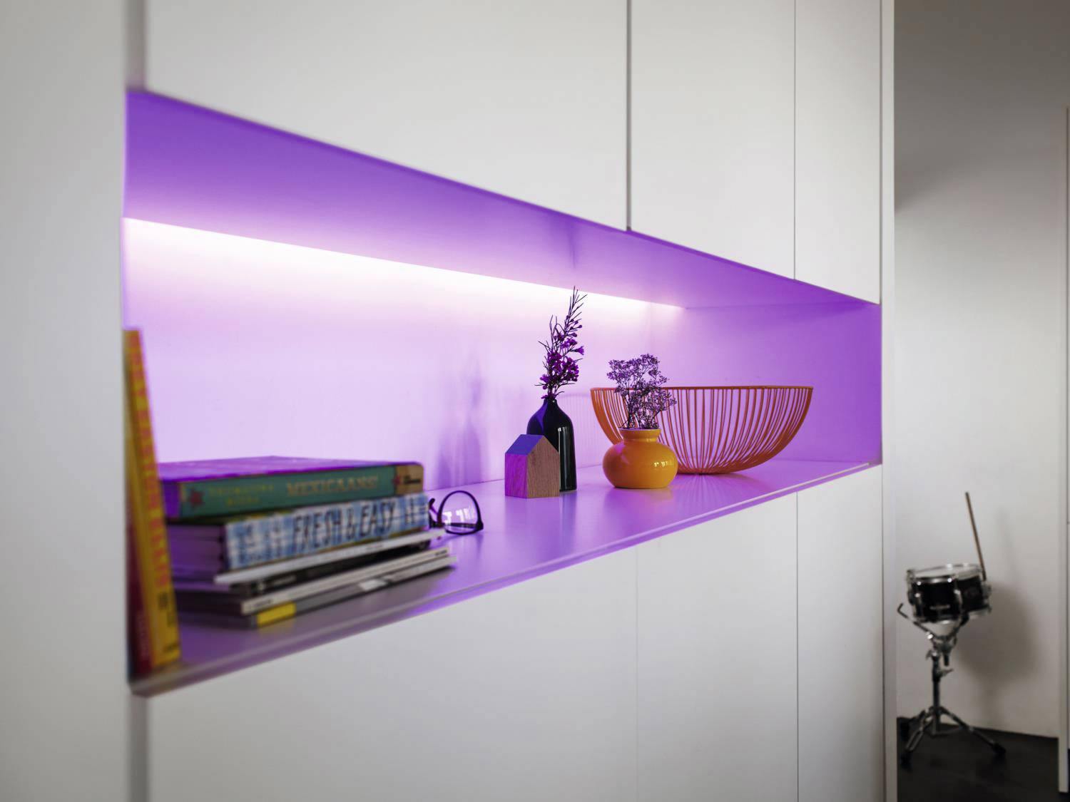 White shelves with lilac backlighting, decorations and books on the left side, an orange basket on the right.