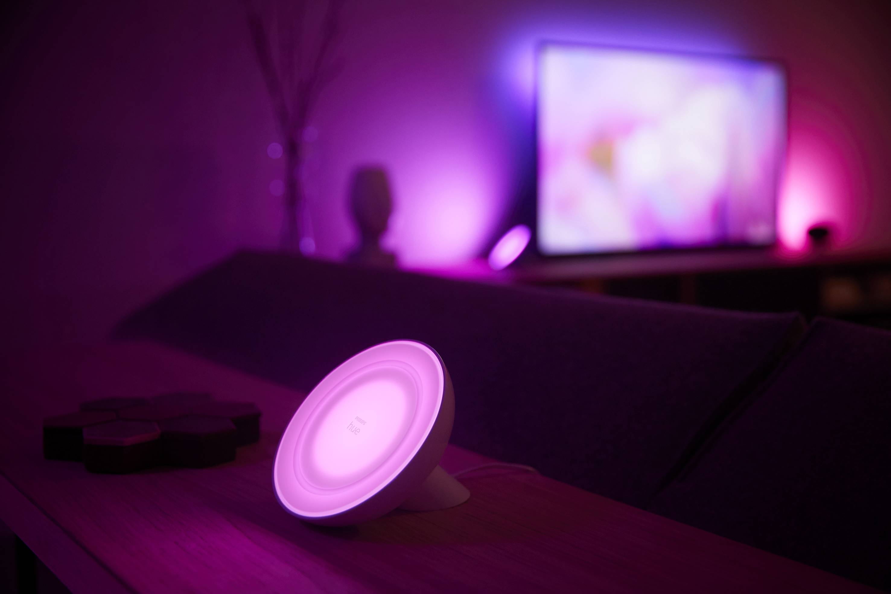 A lamp on a table glows pink, complementing the lights of a television in the background, creating a moody atmosphere.