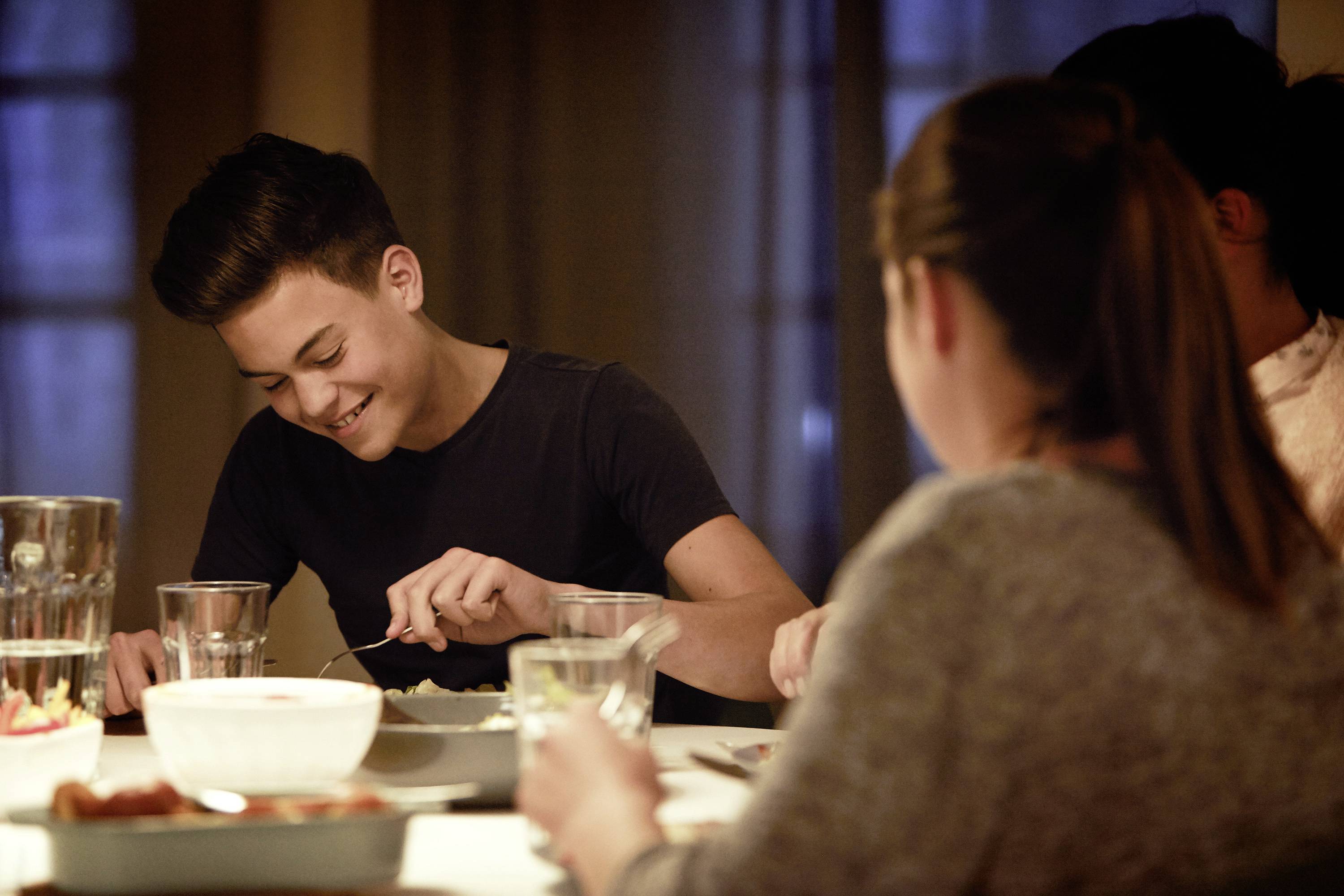 A group of teenagers sits smiling together at a table for dinner. A boy in the foreground is eating in good spirits.