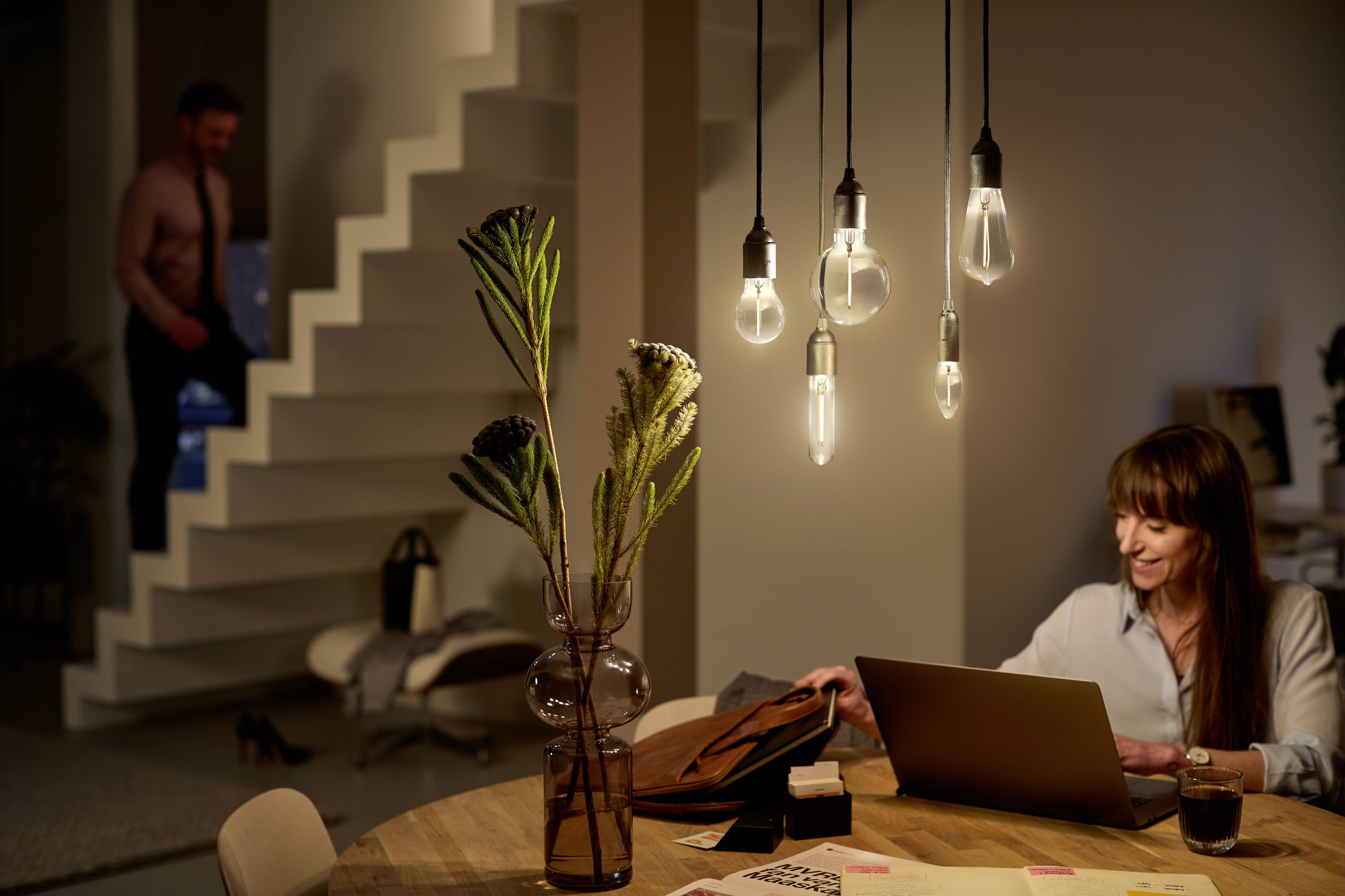 A woman is sitting at a table with a laptop, smiling as she looks at it. In the background, a man is standing on the stairs. The room is warmly lit.