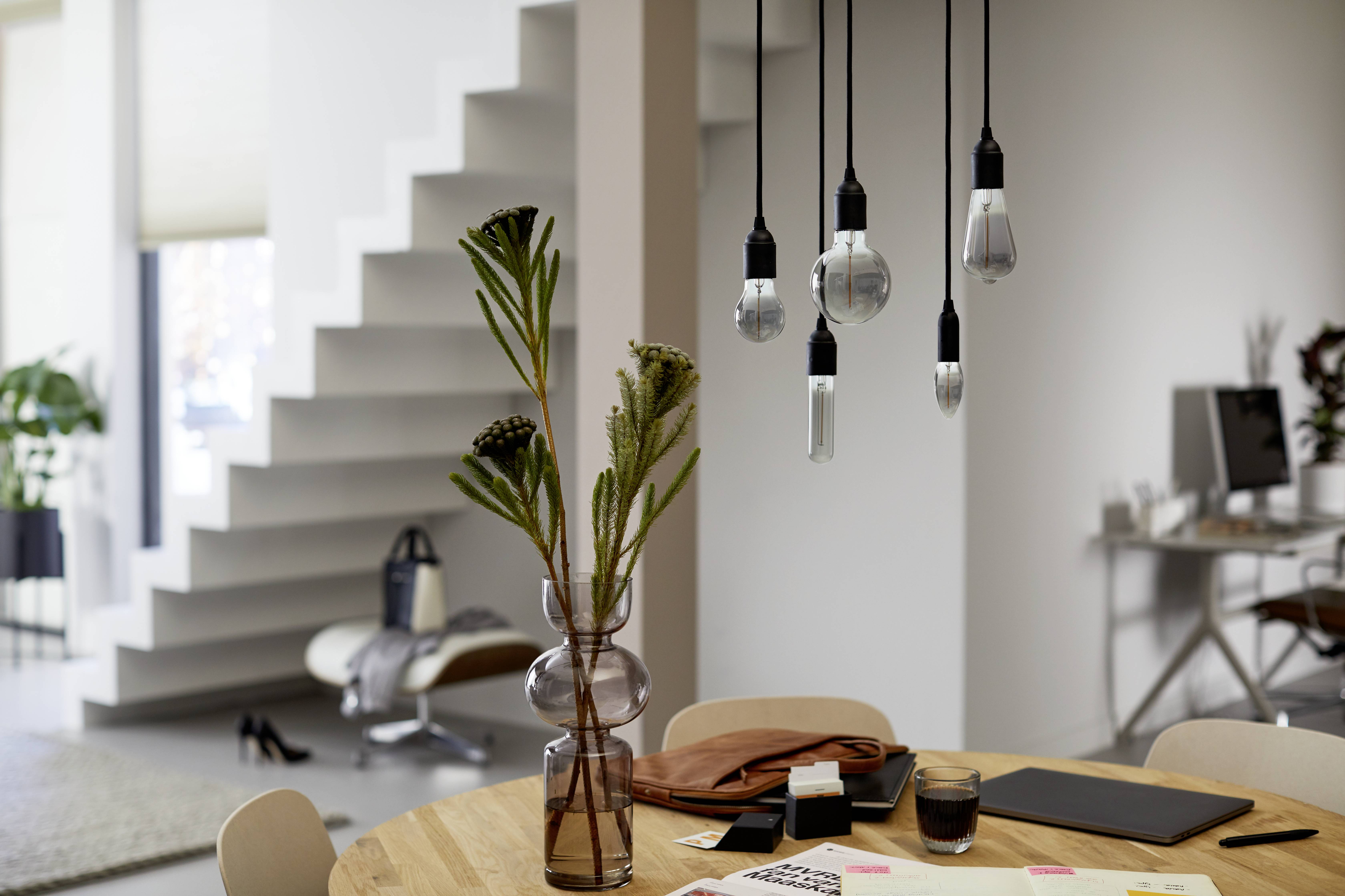 Modern living room with a round wooden table, hanging light bulbs, plant vase and office supplies. Open staircase in the background.