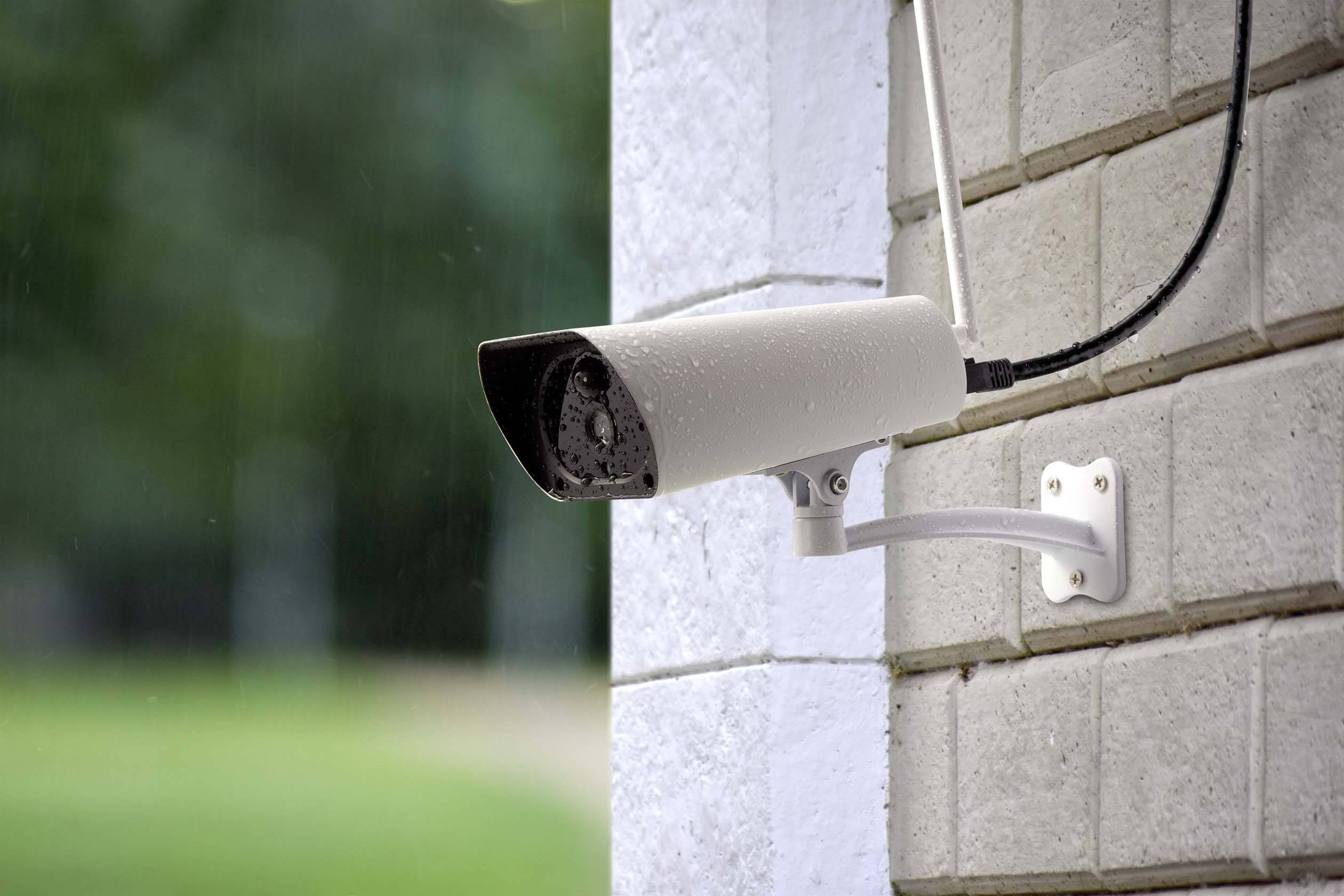 A surveillance camera is mounted on a brick wall. It is facing forward and appears to be operational during light rain.