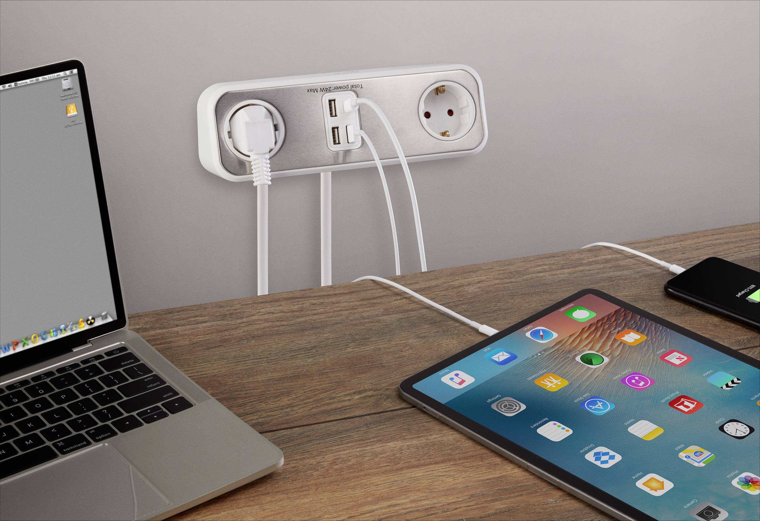 A laptop, tablet and smartphone are connected to a multi-socket outlet on the wall, placed on a table. The laptop has a grey display.