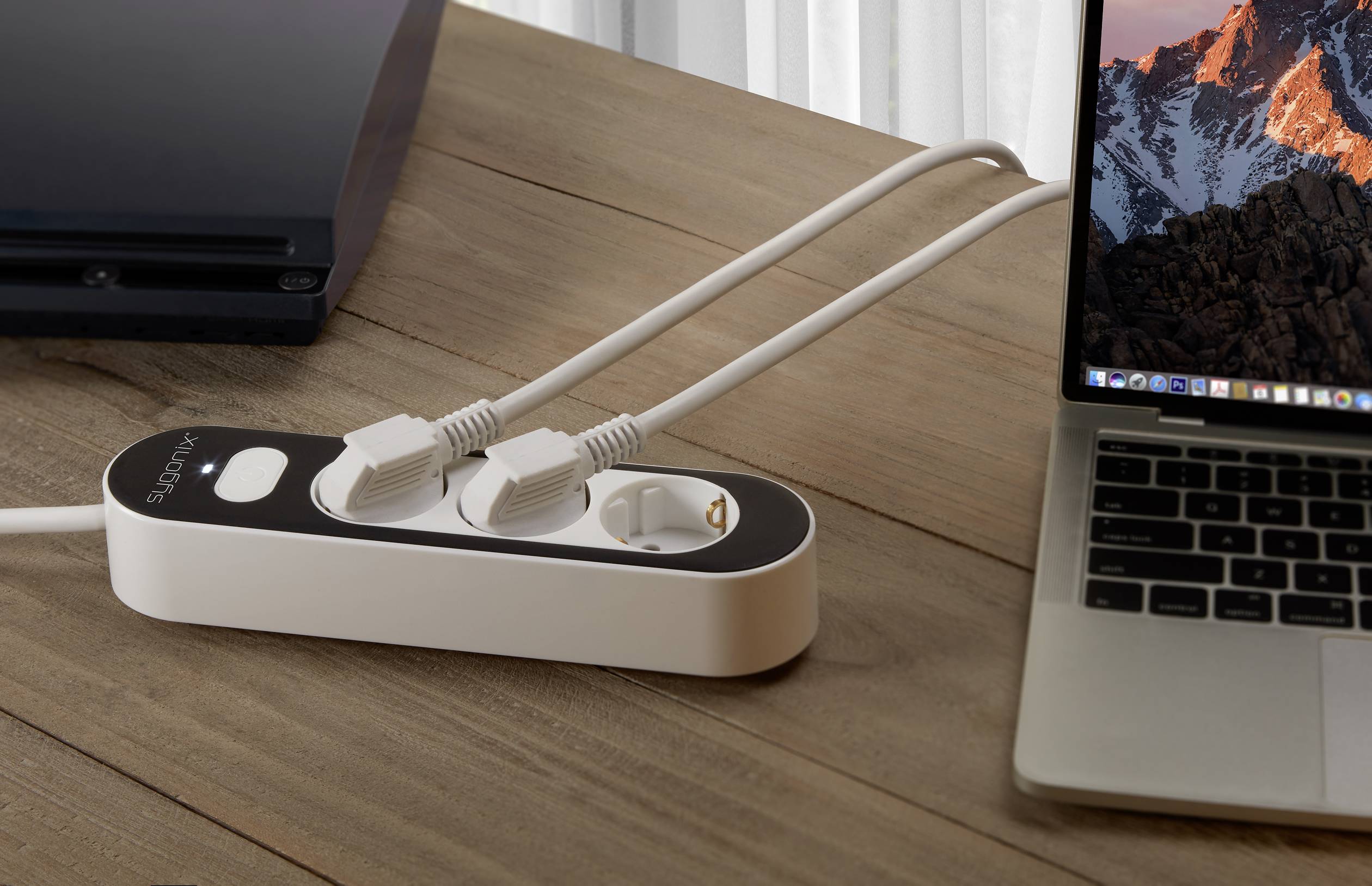 A laptop is connected to a power socket via an extension lead, which is situated on a wooden table. Next to the laptop is a black device.