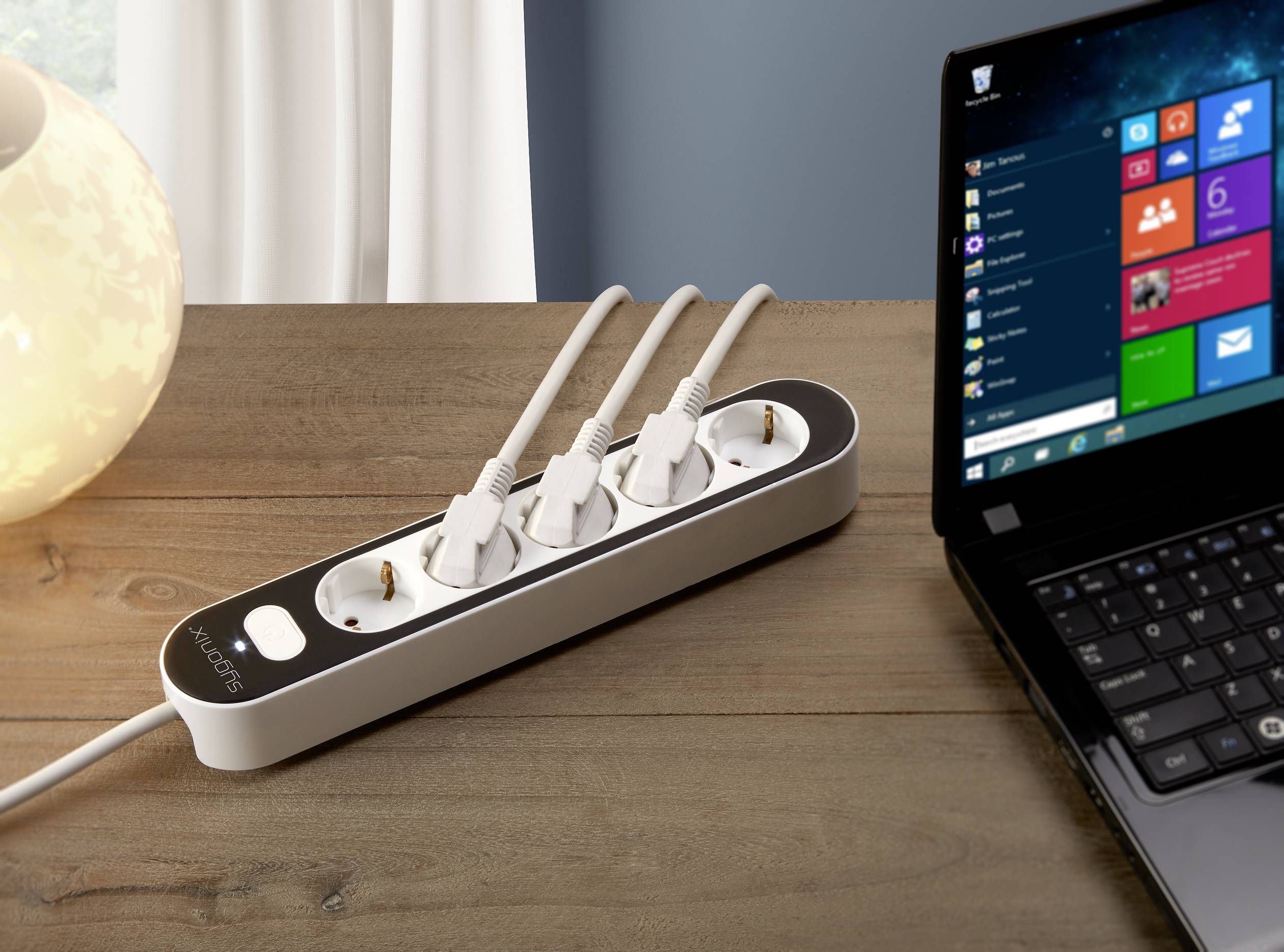 Multi-socket extension lead with three plugged-in cables next to a laptop on a wooden table; a softly illuminated light in the background.
