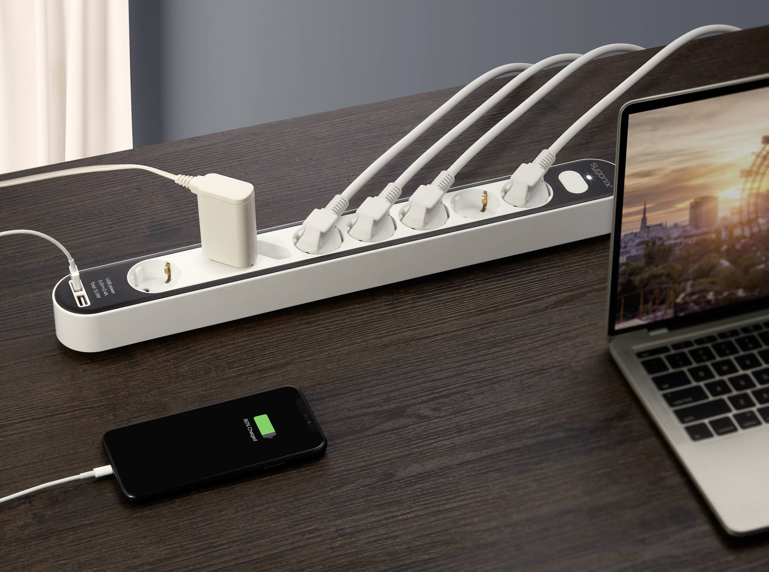 Multiple power socket on a wooden table with five plugged-in cables, smartphone charging, laptop switched on beside it.