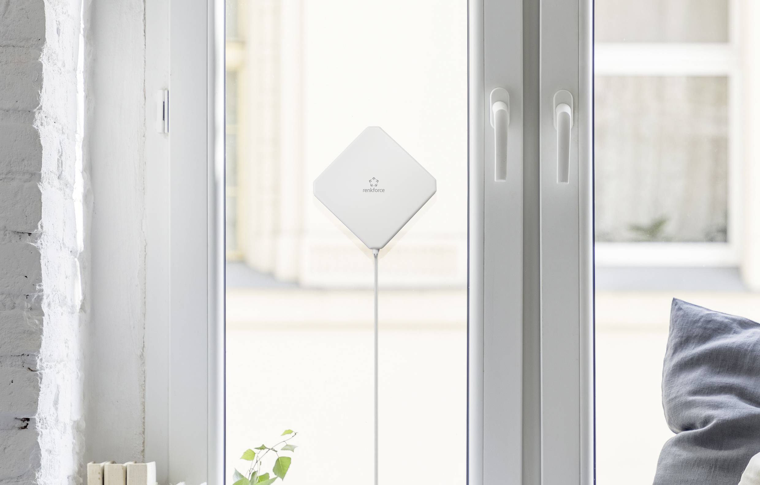 A white technical device stands in front of a large window. It is rectangular and attached to a thin rod. Window shutters are visible in the background.