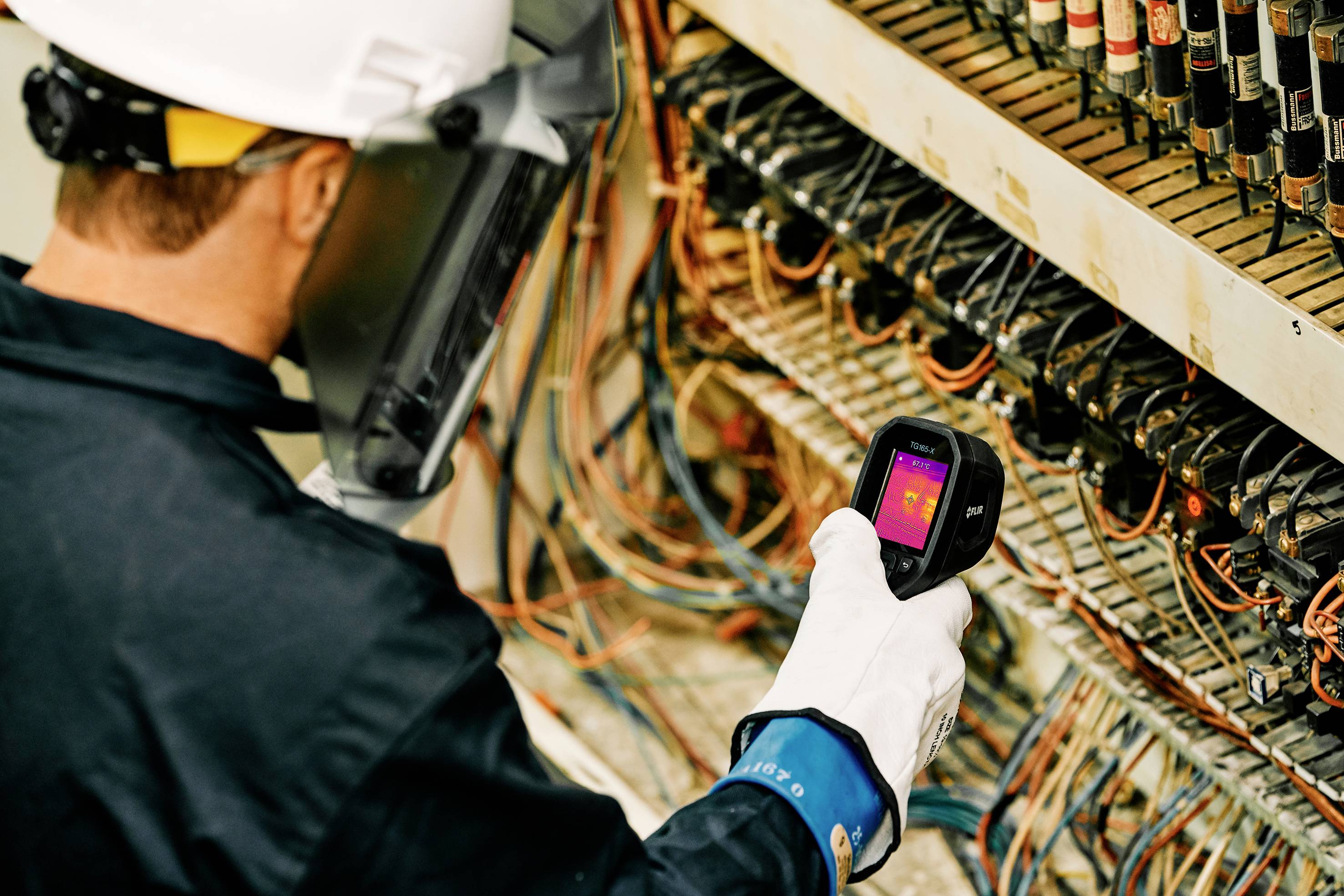 A technician wearing protective equipment is checking cable connections in a control cabinet using an infrared thermometer.