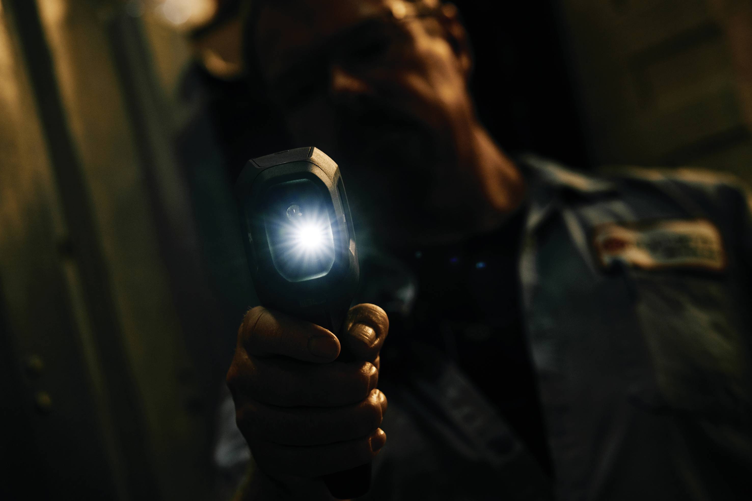 A person is holding an infrared thermometer in a dark environment. The display area of the device is illuminated, while the surrounding area remains in shadow.
