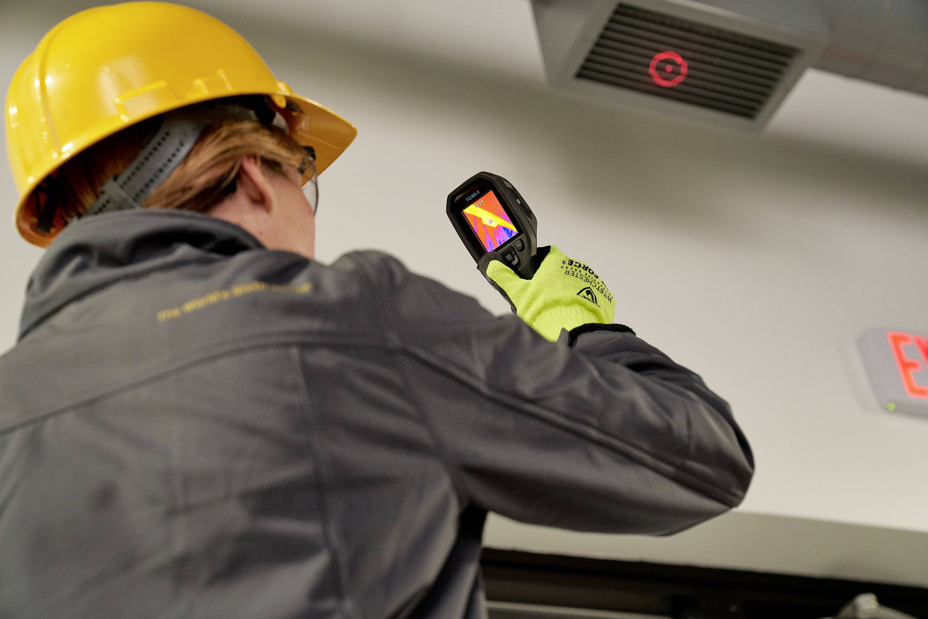A person wearing a protective helmet and gloves is using a thermal imaging camera to check the temperature on a ventilation unit.