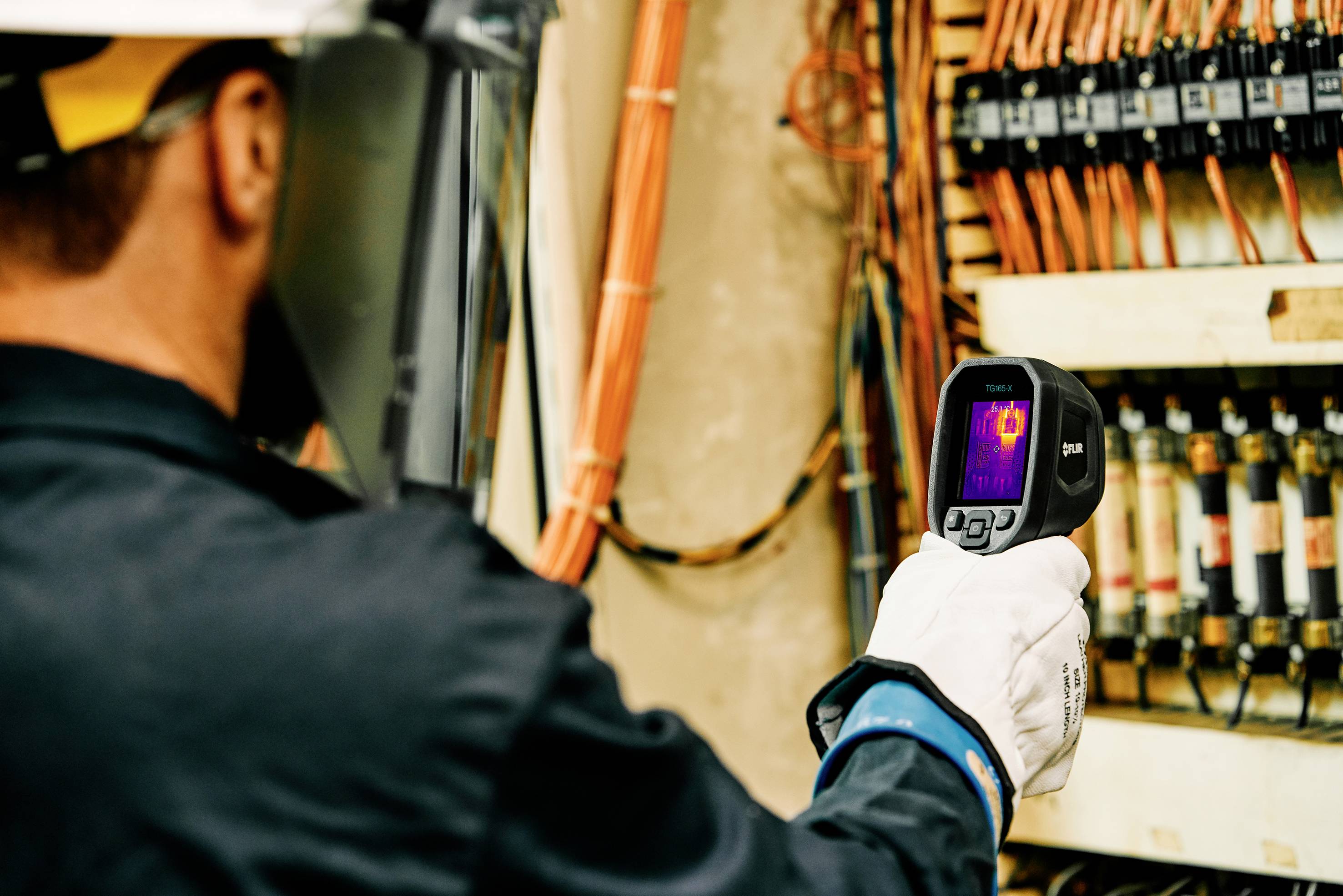 A technician in safety equipment is using a thermal imaging camera to check the temperatures of electrical cables.