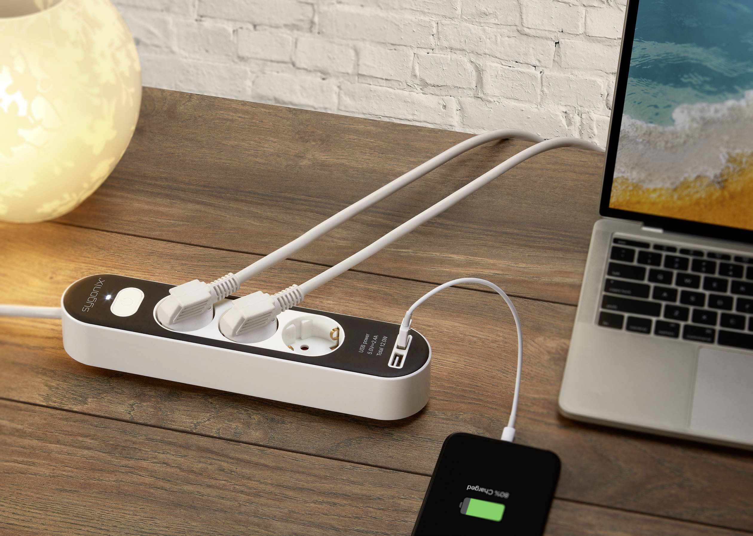 A power strip on a wooden table supplies a laptop and a smartphone with electricity. The smartphone displays a charging process.