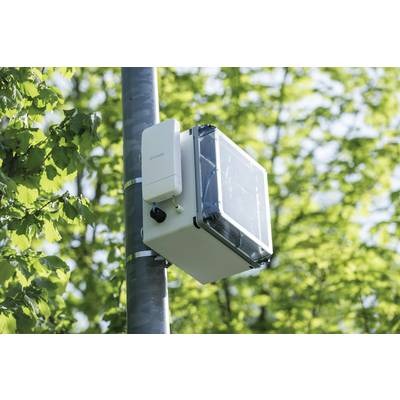 A weather station with a sensor and a solar panel box is attached to a pole. Background: green leaves.