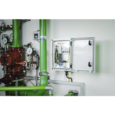 An open electrical cabinet in an industrial space reveals visible cables and wiring. Green pipework and measuring instruments surround the cabinet.