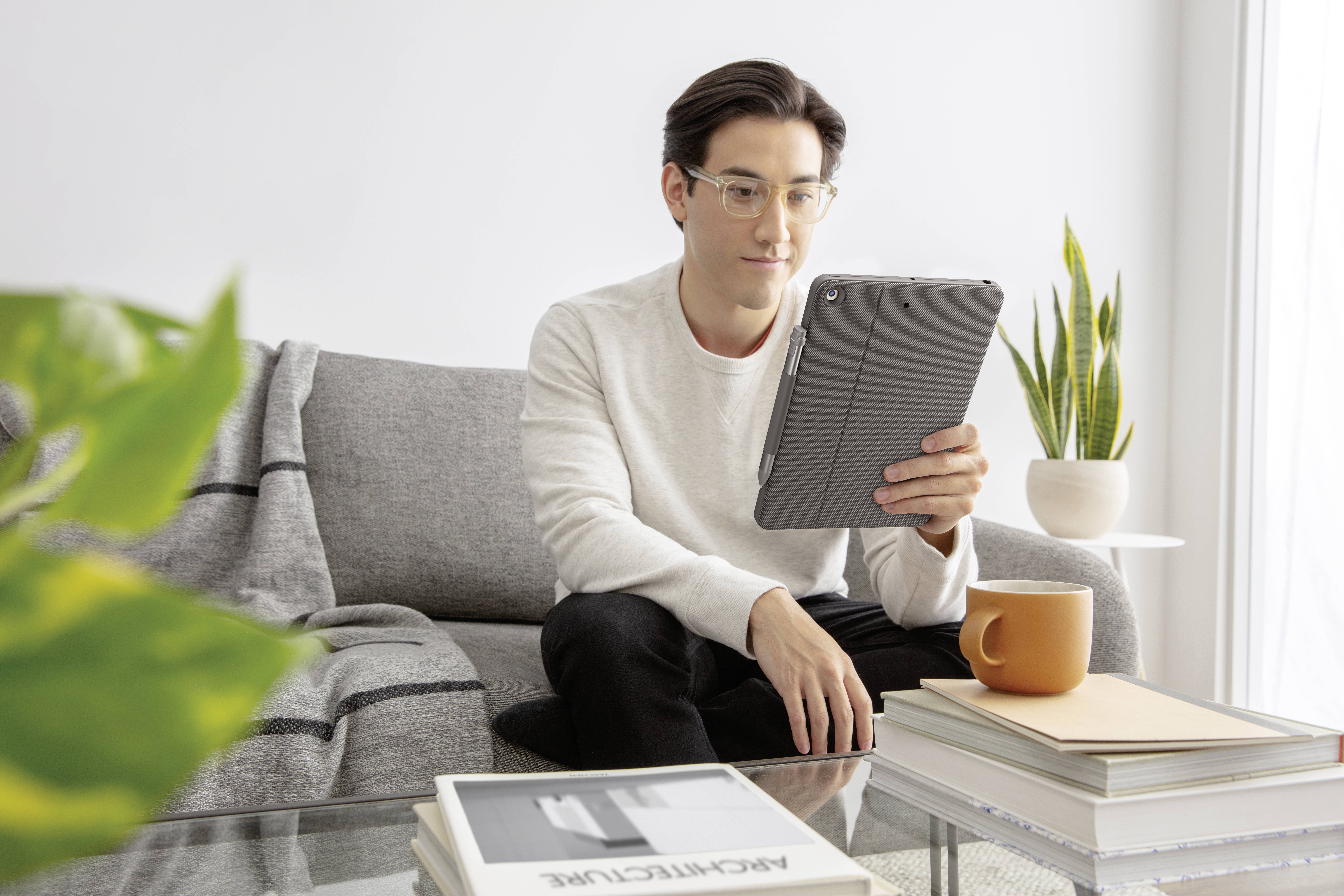 A person is sitting on a sofa and reading on a tablet. A table with books, a mug, and plants is in the foreground.