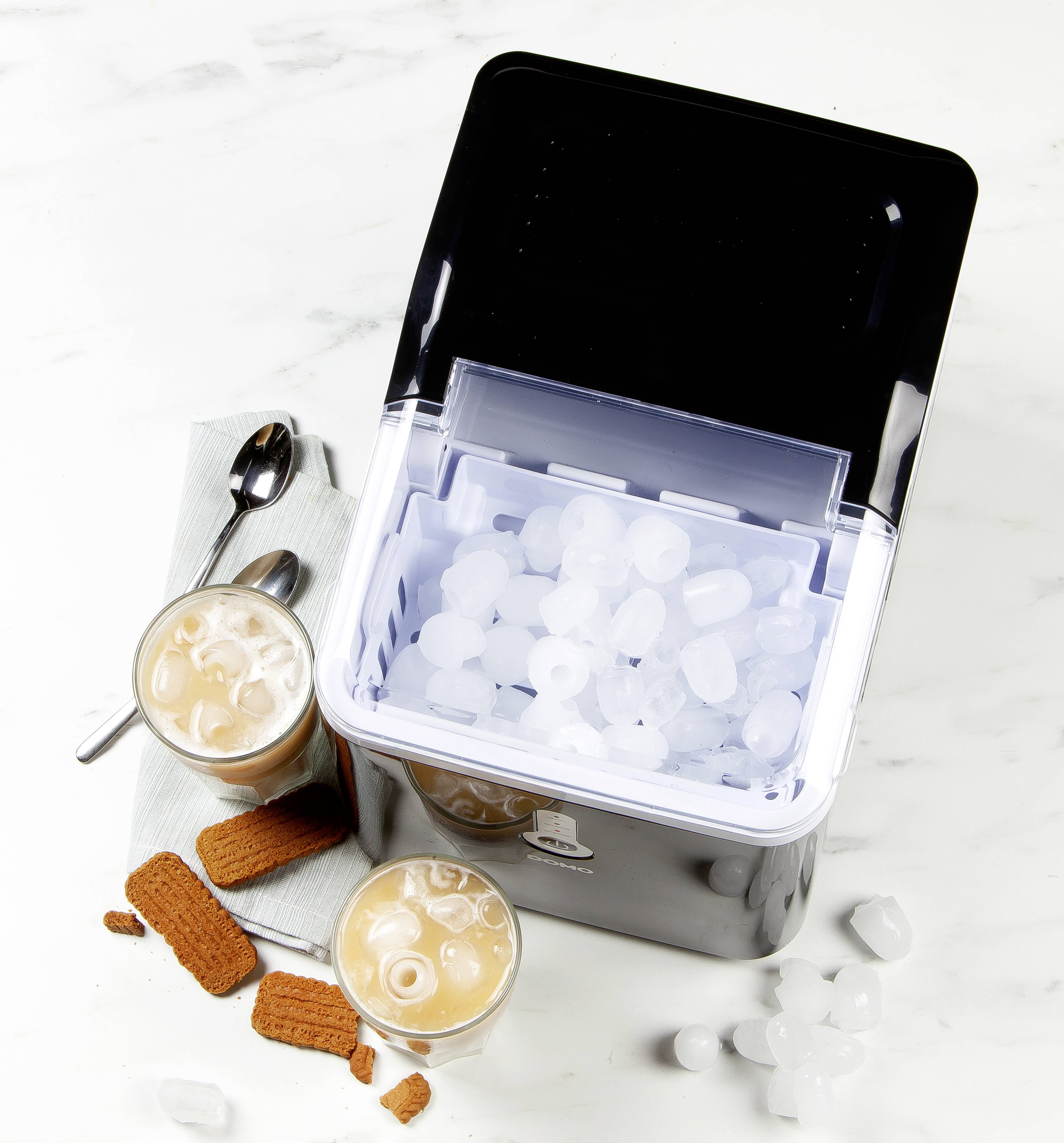 A black ice maker, filled with round ice cubes, sits beside two glasses of iced coffee and biscuits on a light surface.