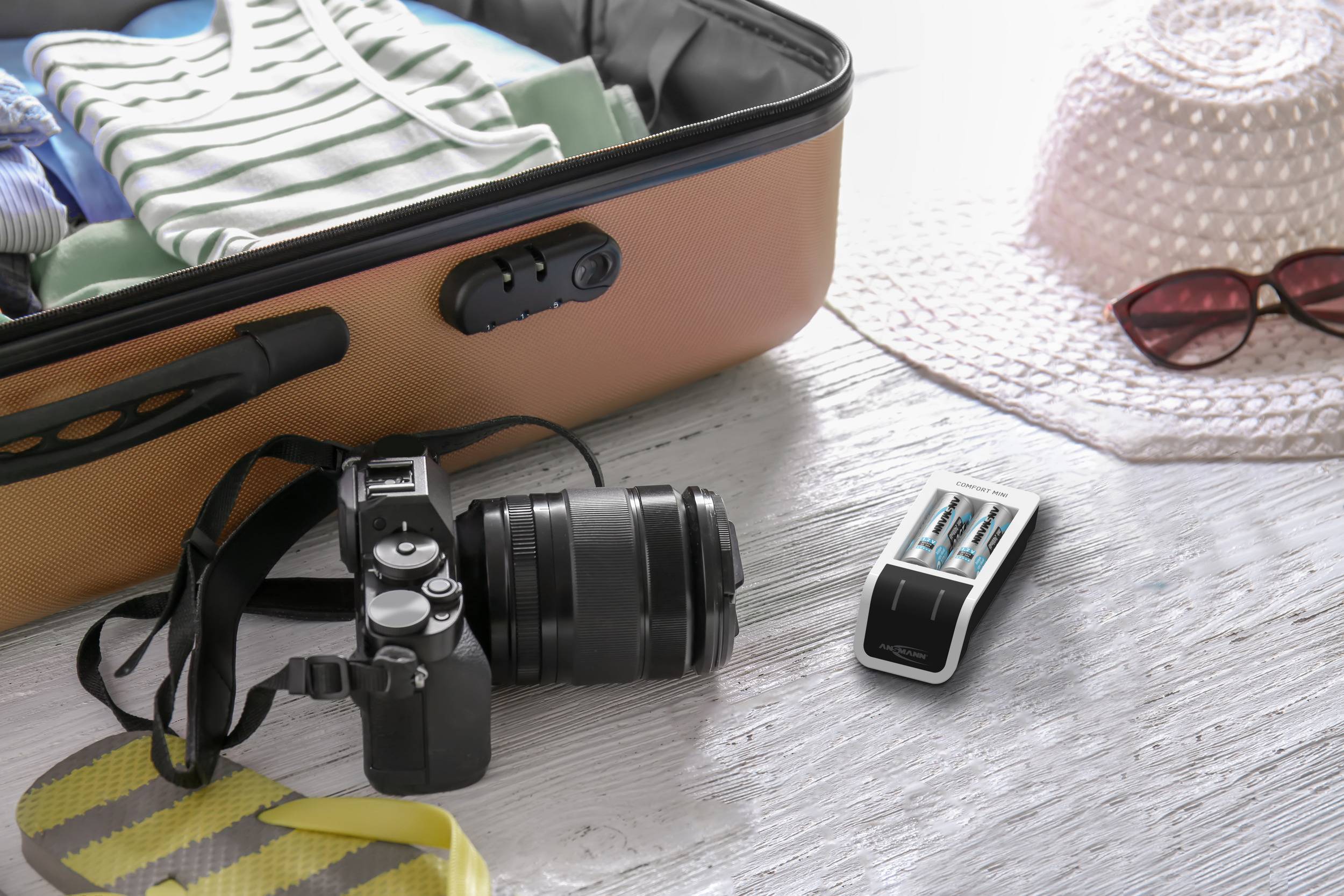 Open suitcase with clothing, camera and battery charger on light wooden table, with a sun hat and sunglasses beside it. Travel preparation.