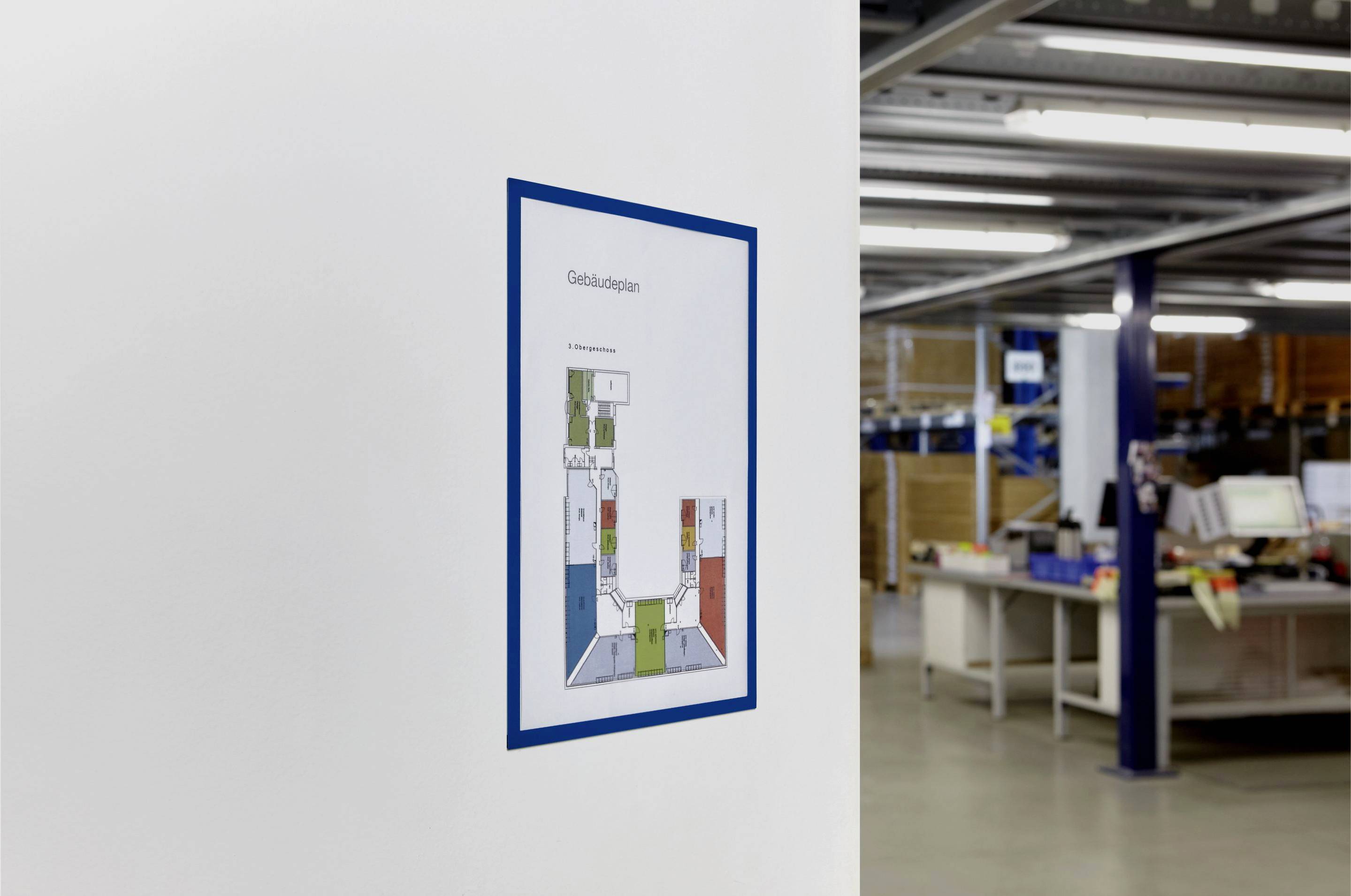 A building plan hangs on a white wall in an office area with tables and shelves in the background.