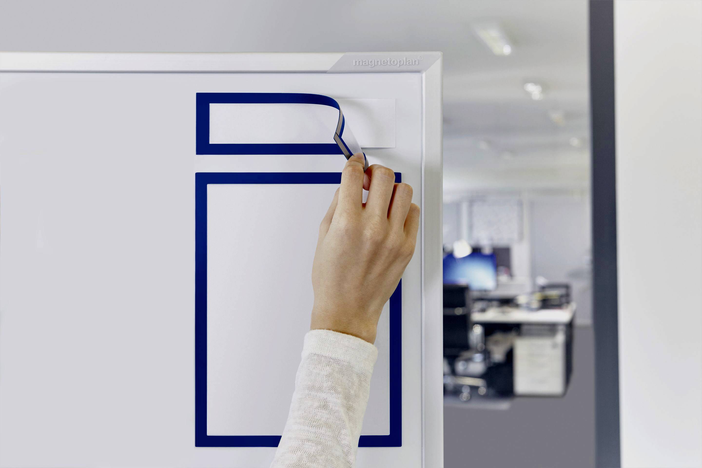 A hand sticks a piece of paper to a board in a modern office. Office furniture and computers are blurred in the background.
