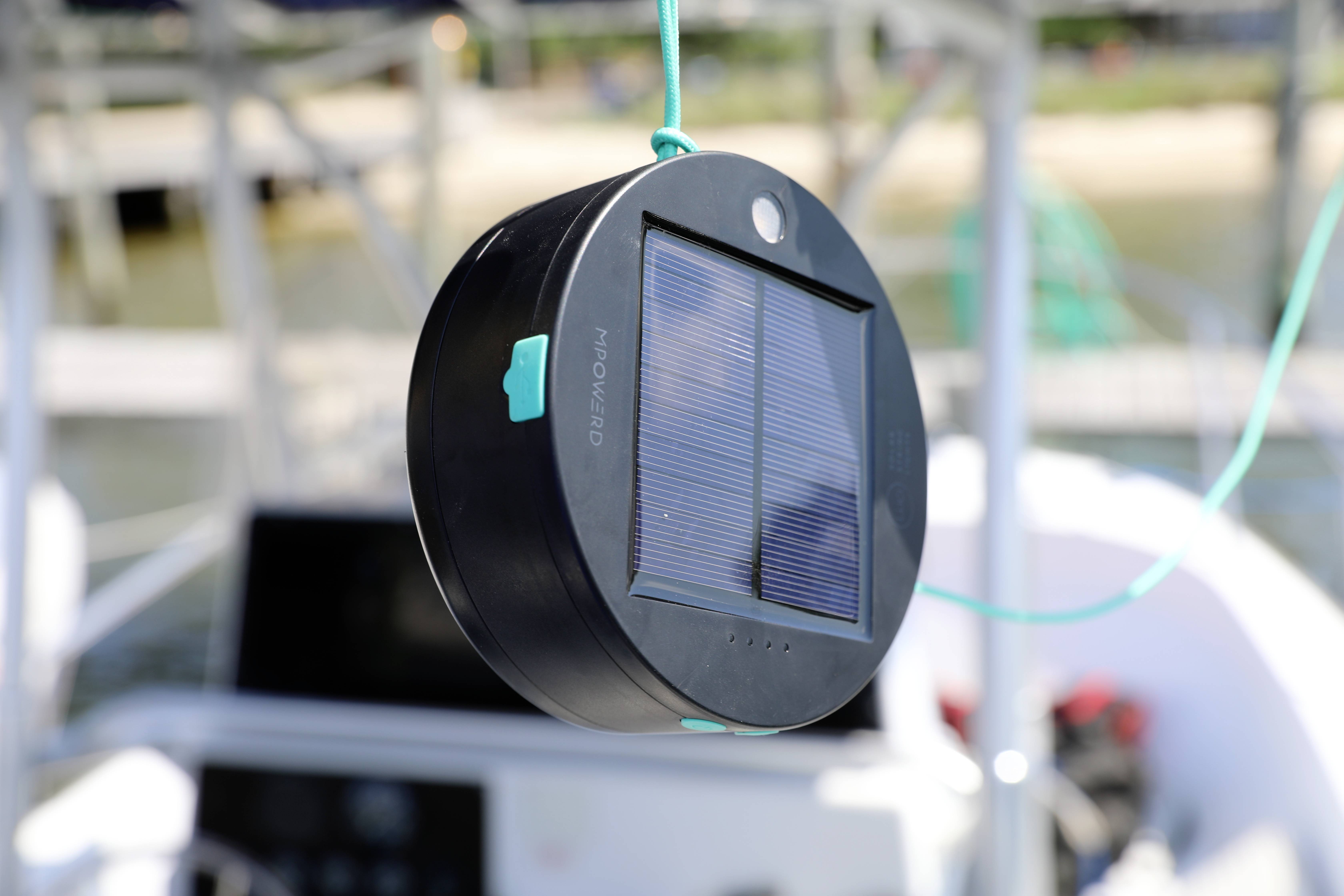 A portable solar lamp with a solar panel hangs from a rope in an outdoor area. Blurred objects can be seen in the background.