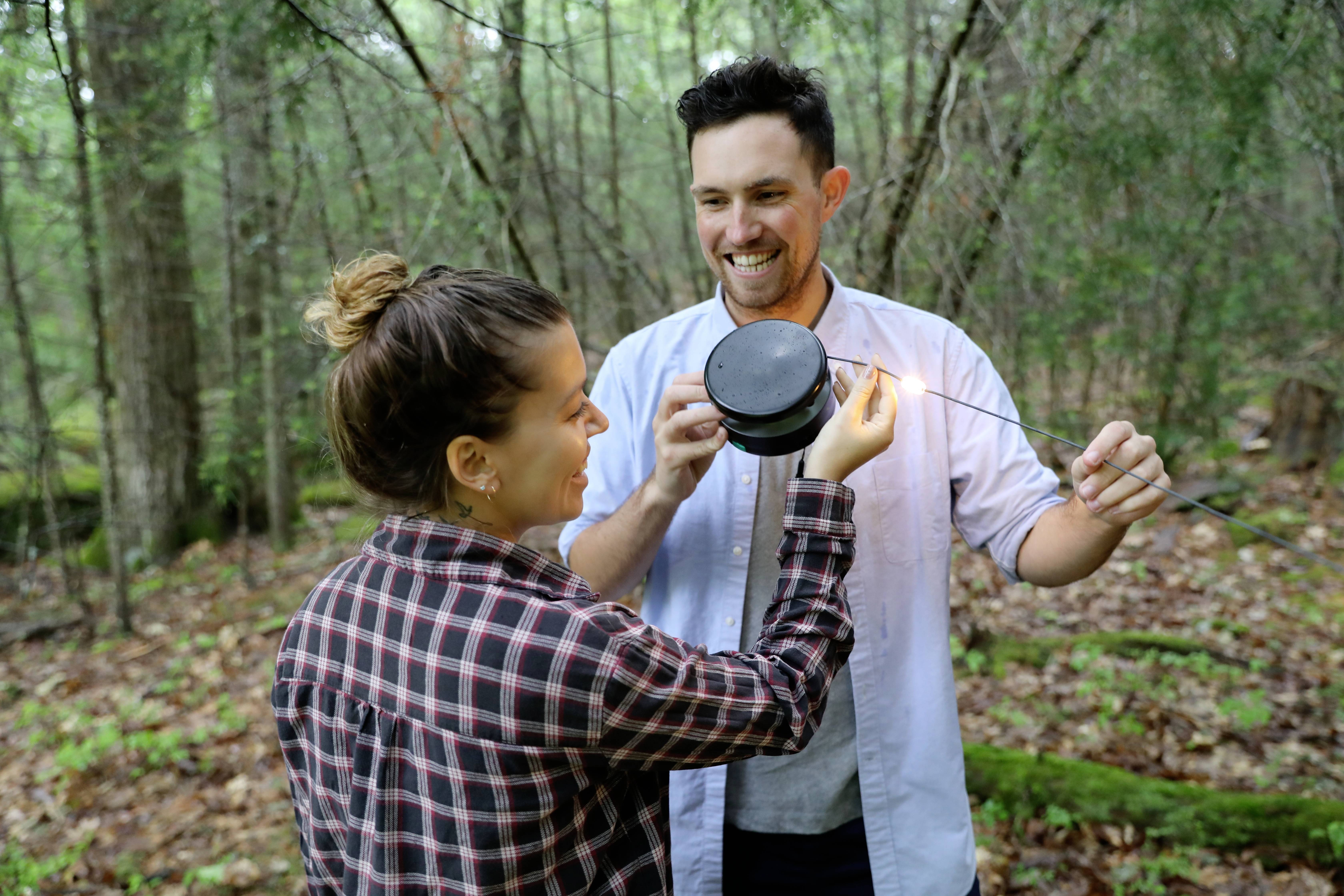 A man and a woman are standing in the woods and working together on a camera. They are smiling and surrounded by trees.