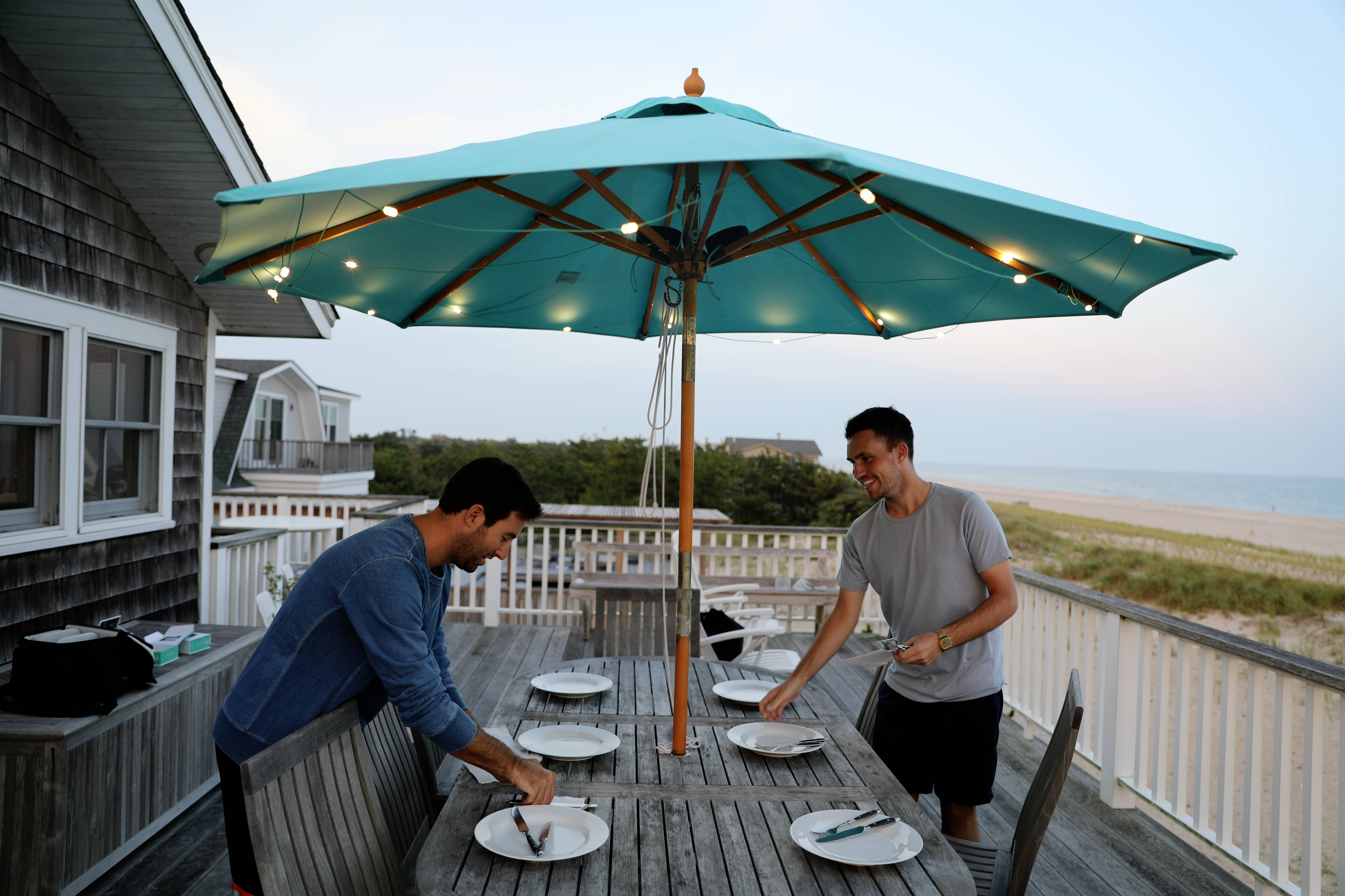 Two people are setting a dining table under a turquoise parasol on a terrace by the beach.