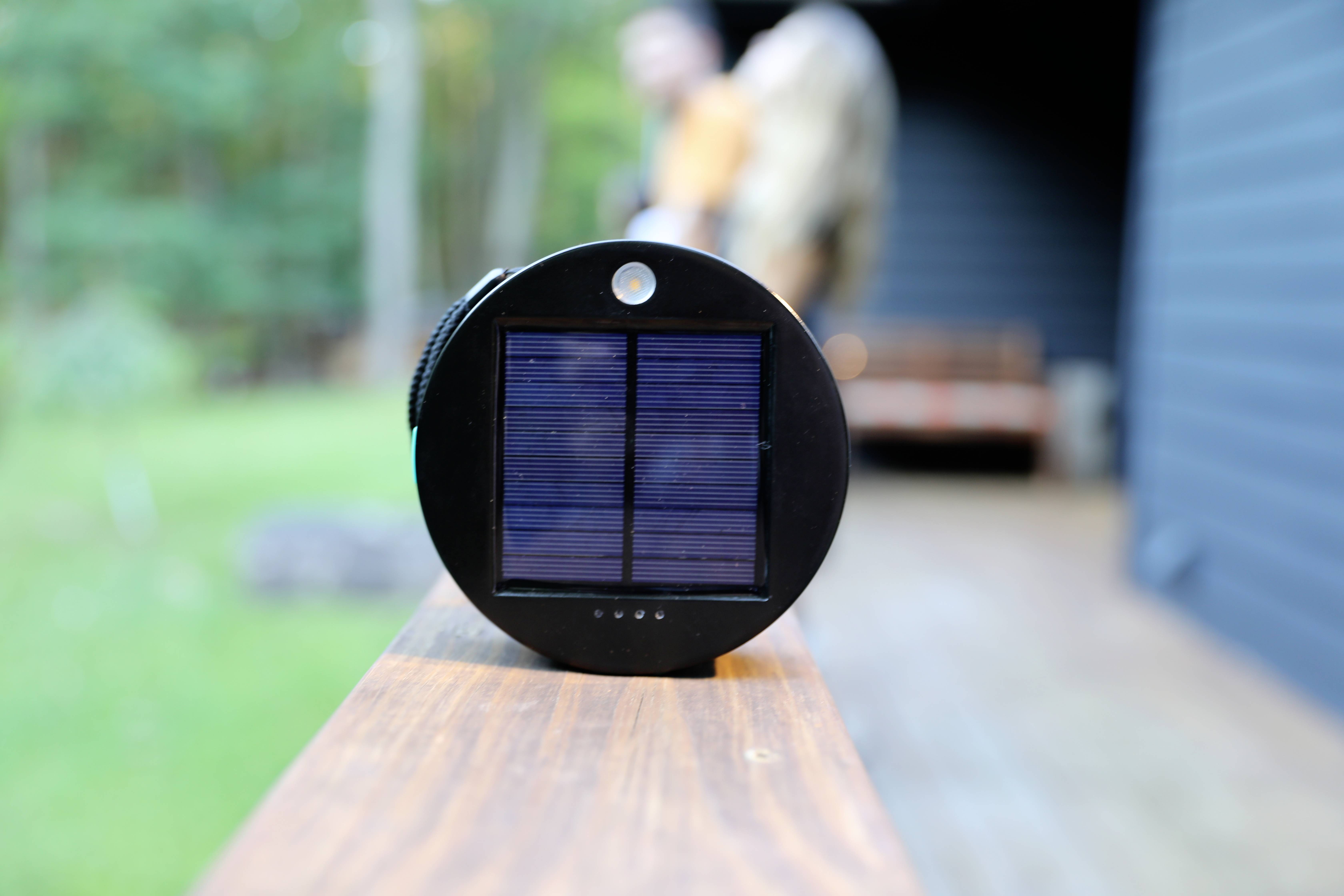 A round solar charger rests on a wooden beam. Trees are visible in the background, with a blurred human silhouette discernible.
