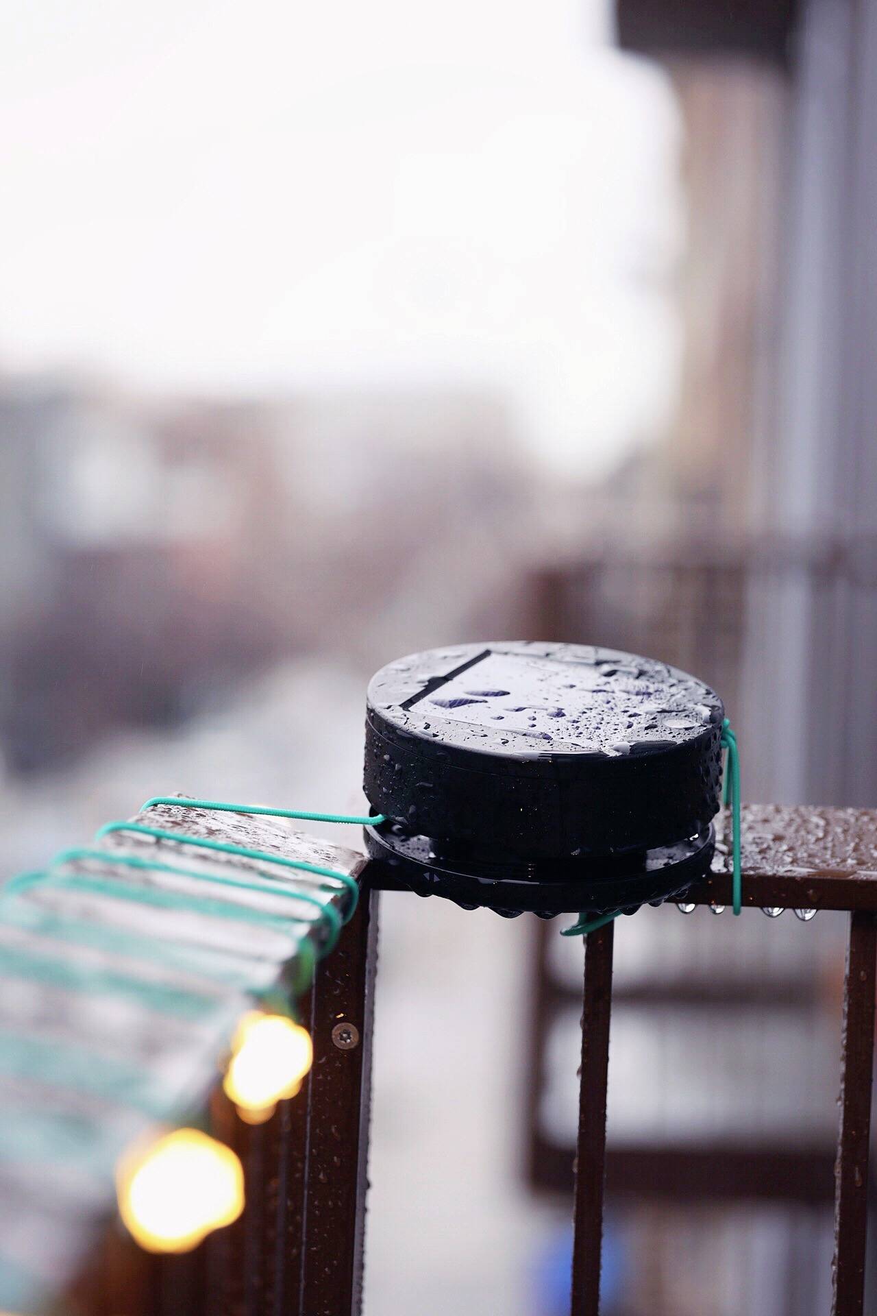 A black speaker sits on a wet balcony railing, with glowing fairy lights hanging beside it. Background blurred.