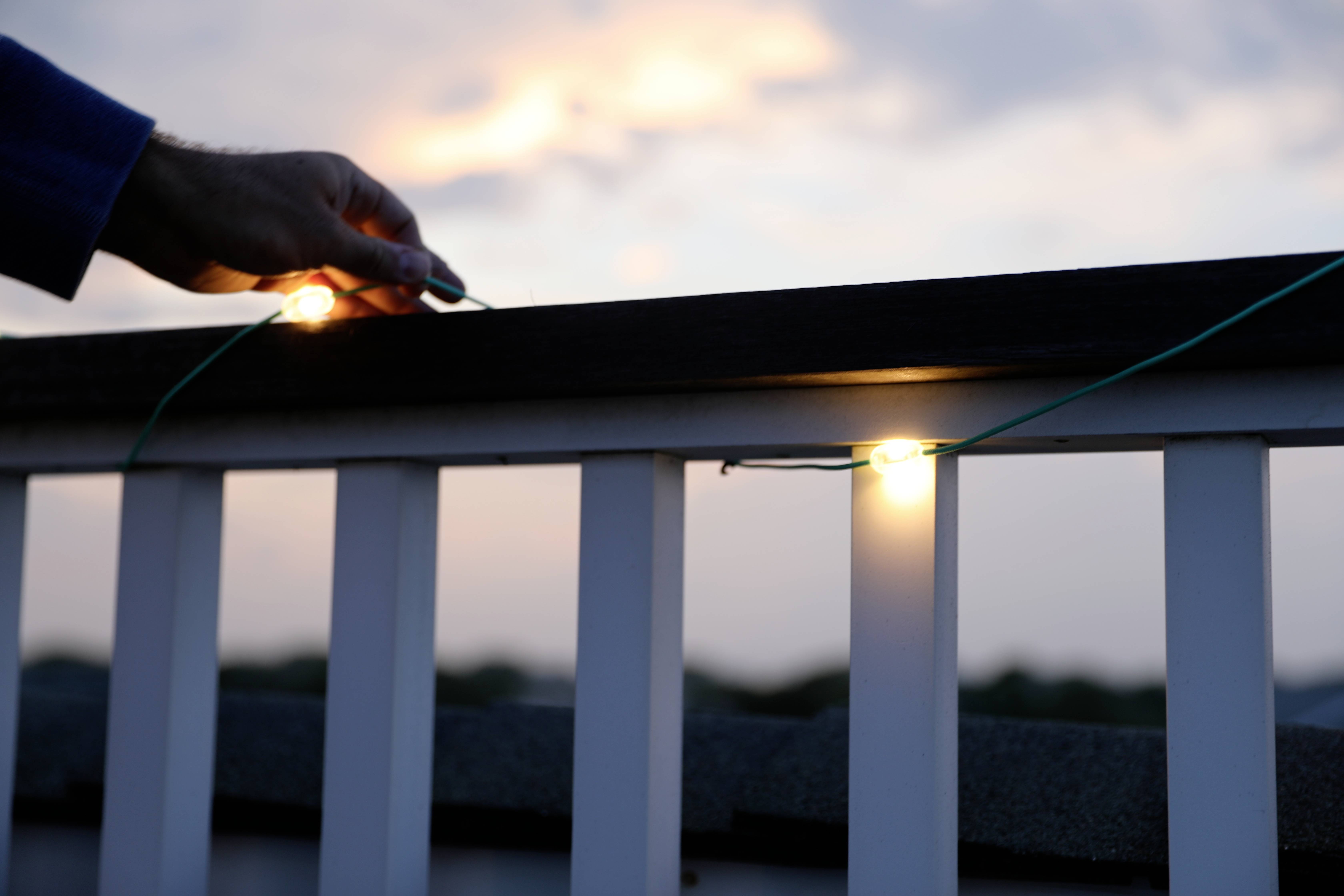 A hand hangs small, glowing lights on a banister. The sky in the background is slightly cloudy and shows a sunset.