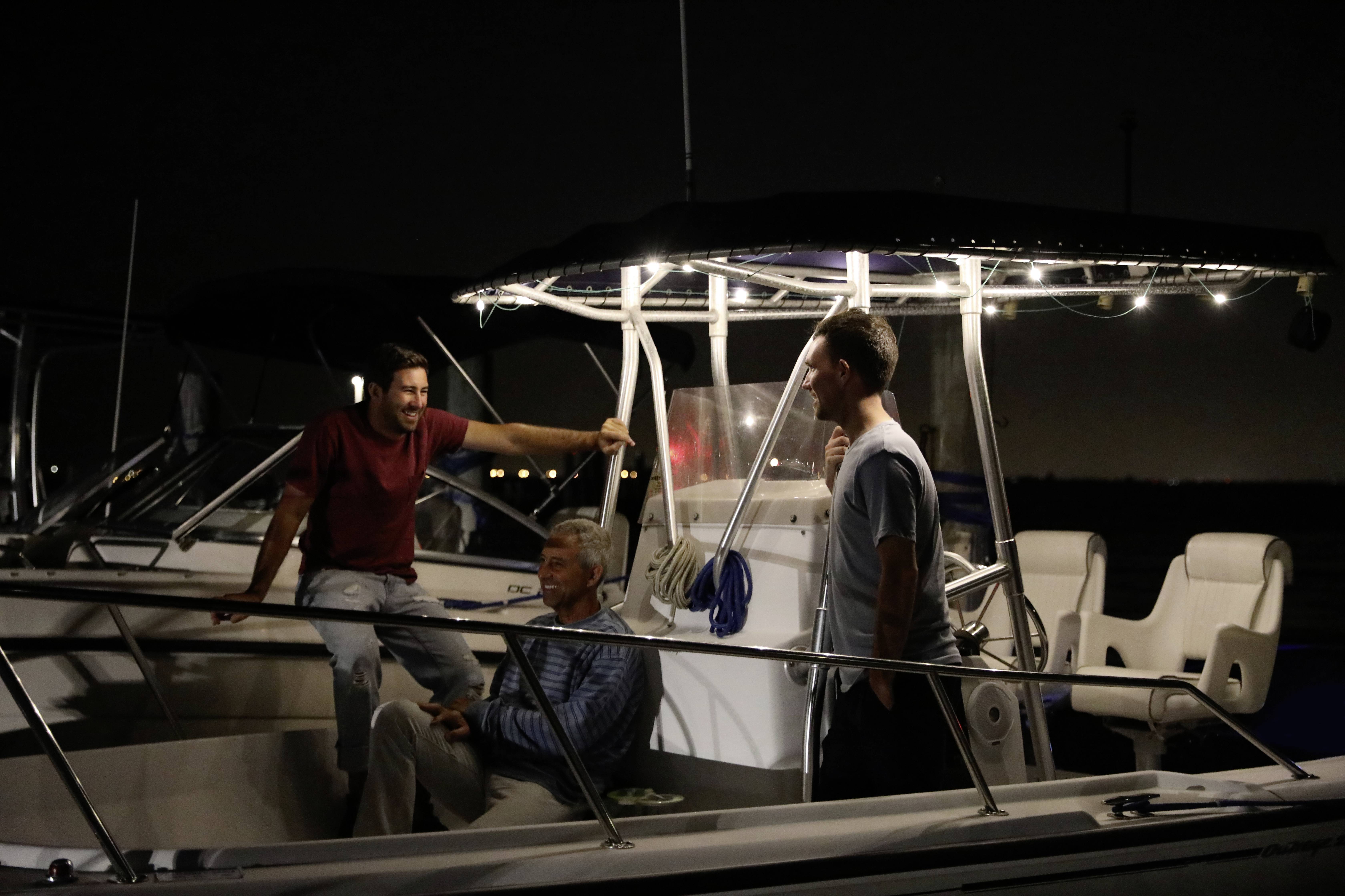 Three people sit relaxed on a boat at night, under soft lighting, and are chatting with one another.