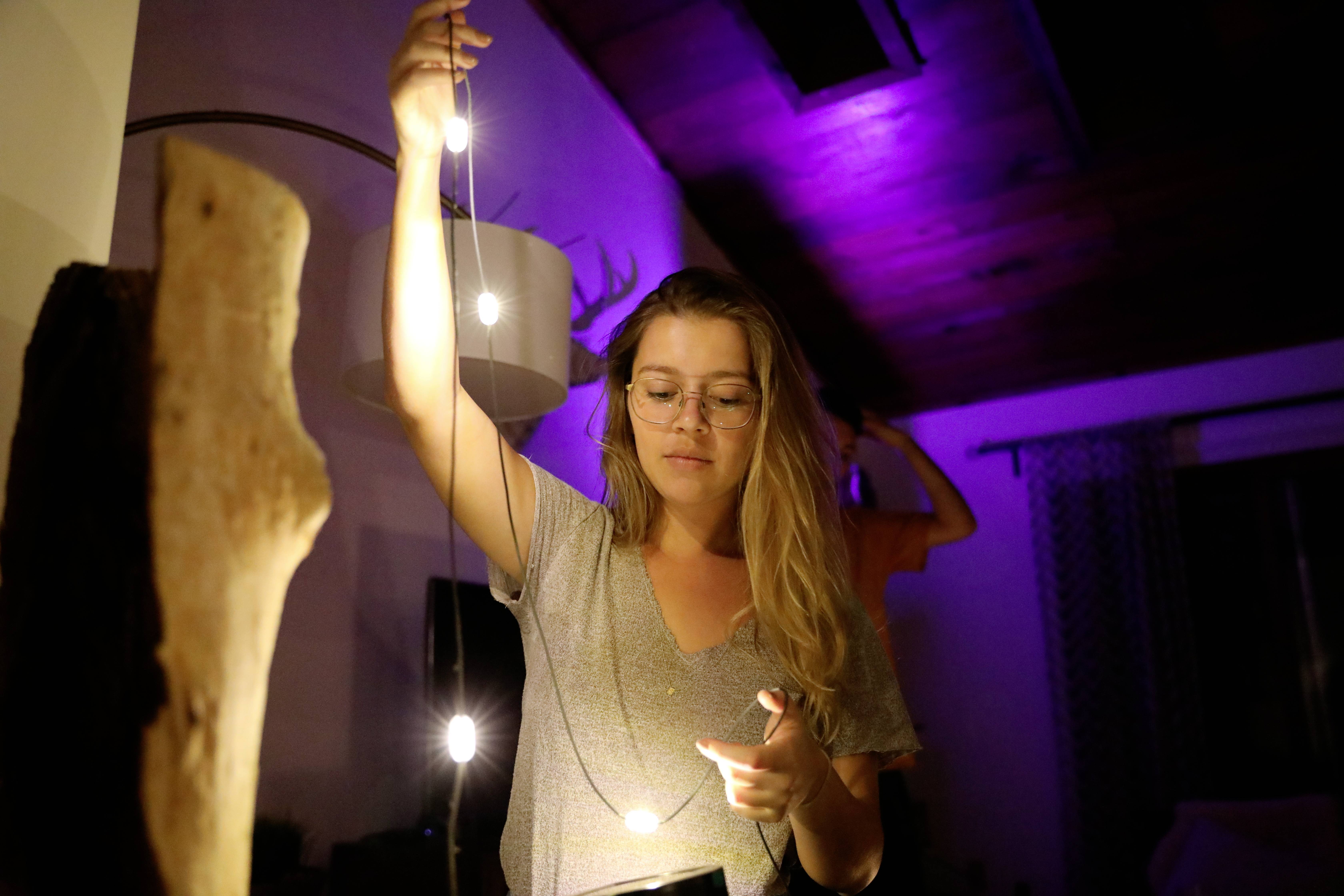 A woman in an illuminated room is holding a string of fairy lights. The background is bathed in purple light. A second person is out of focus.