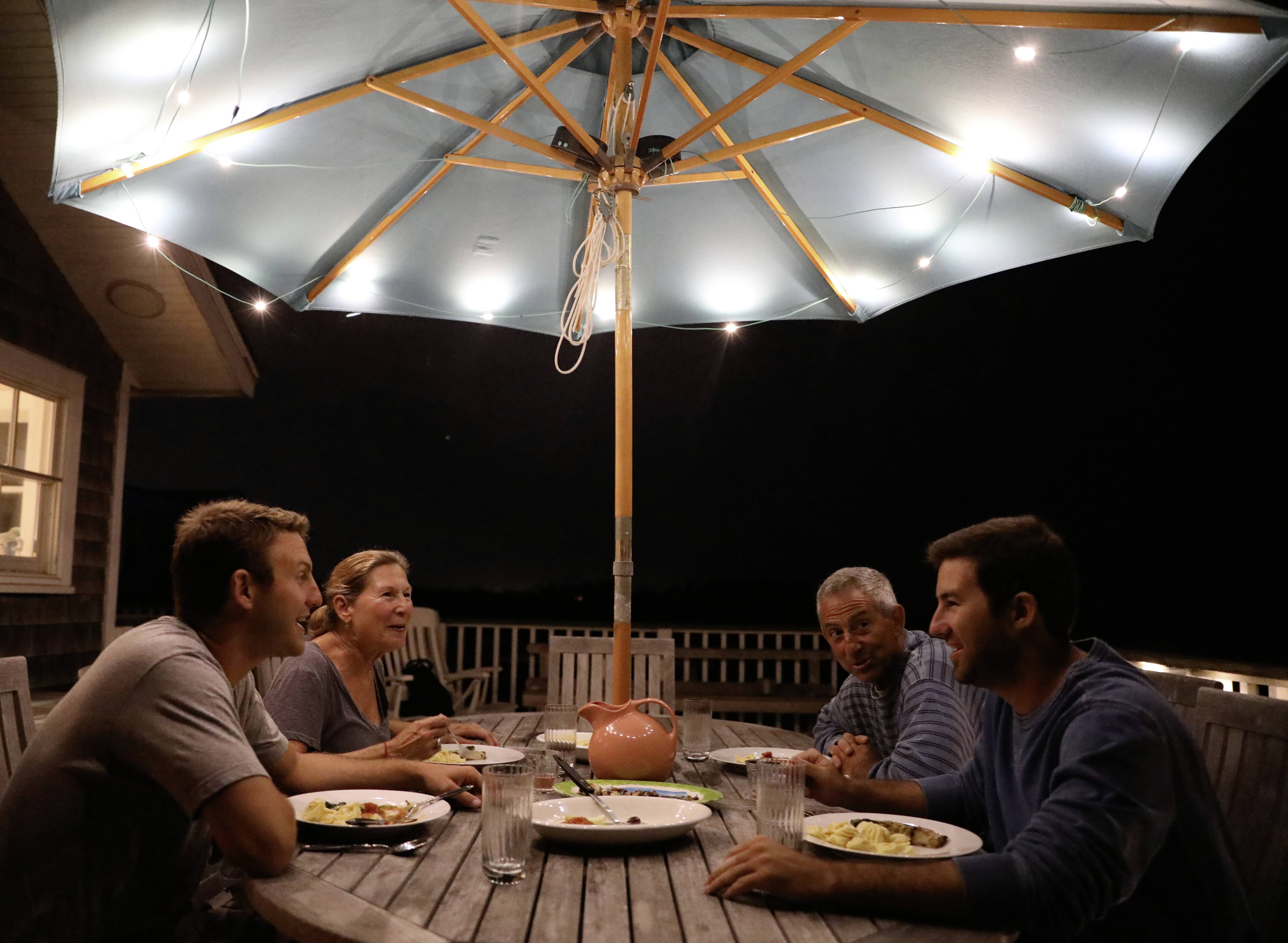 A group of four people are sitting under a lit parasol at an outdoor table at night, eating together.