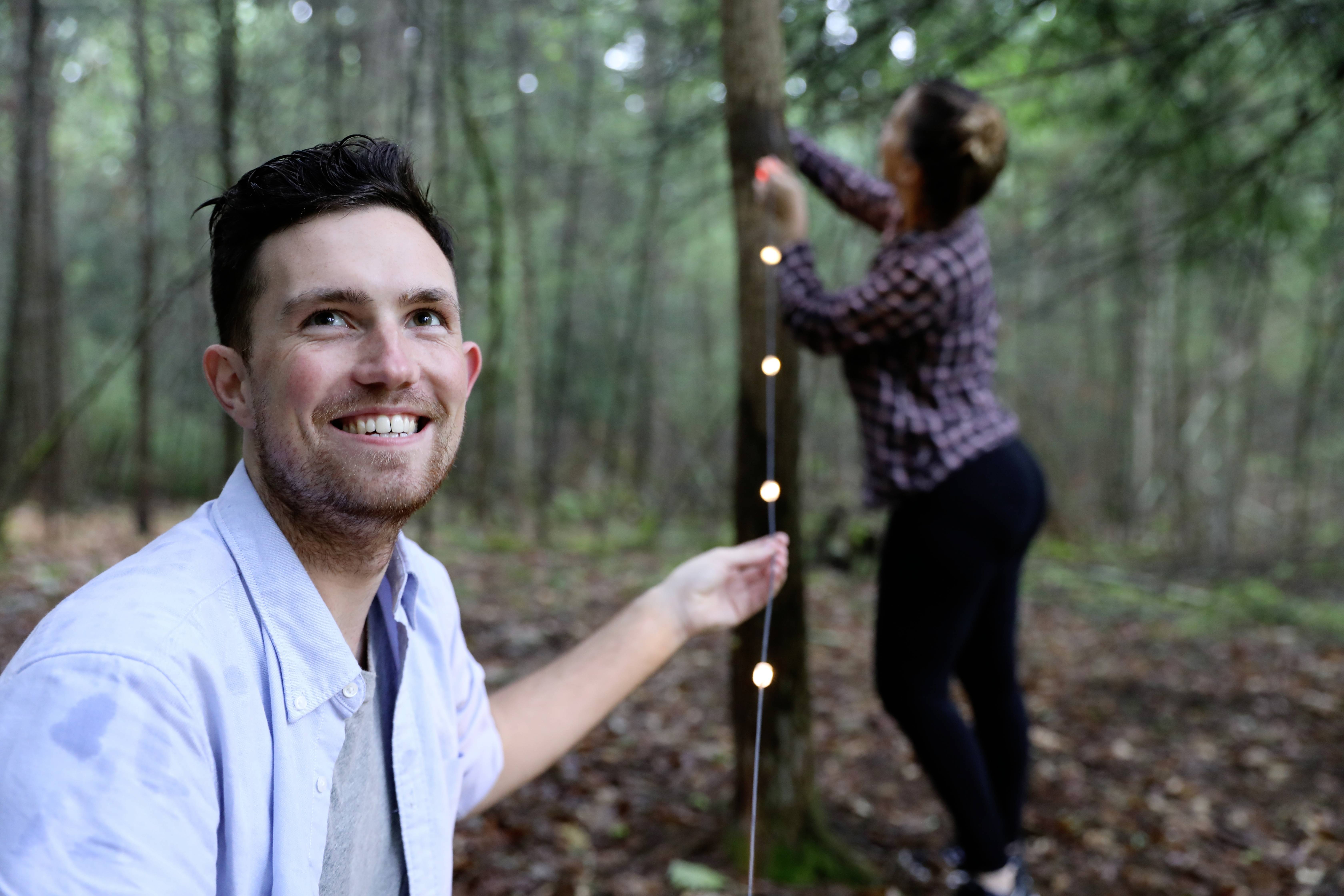 A man is smiling at the camera while another person hangs fairy lights on a tree in the forest.