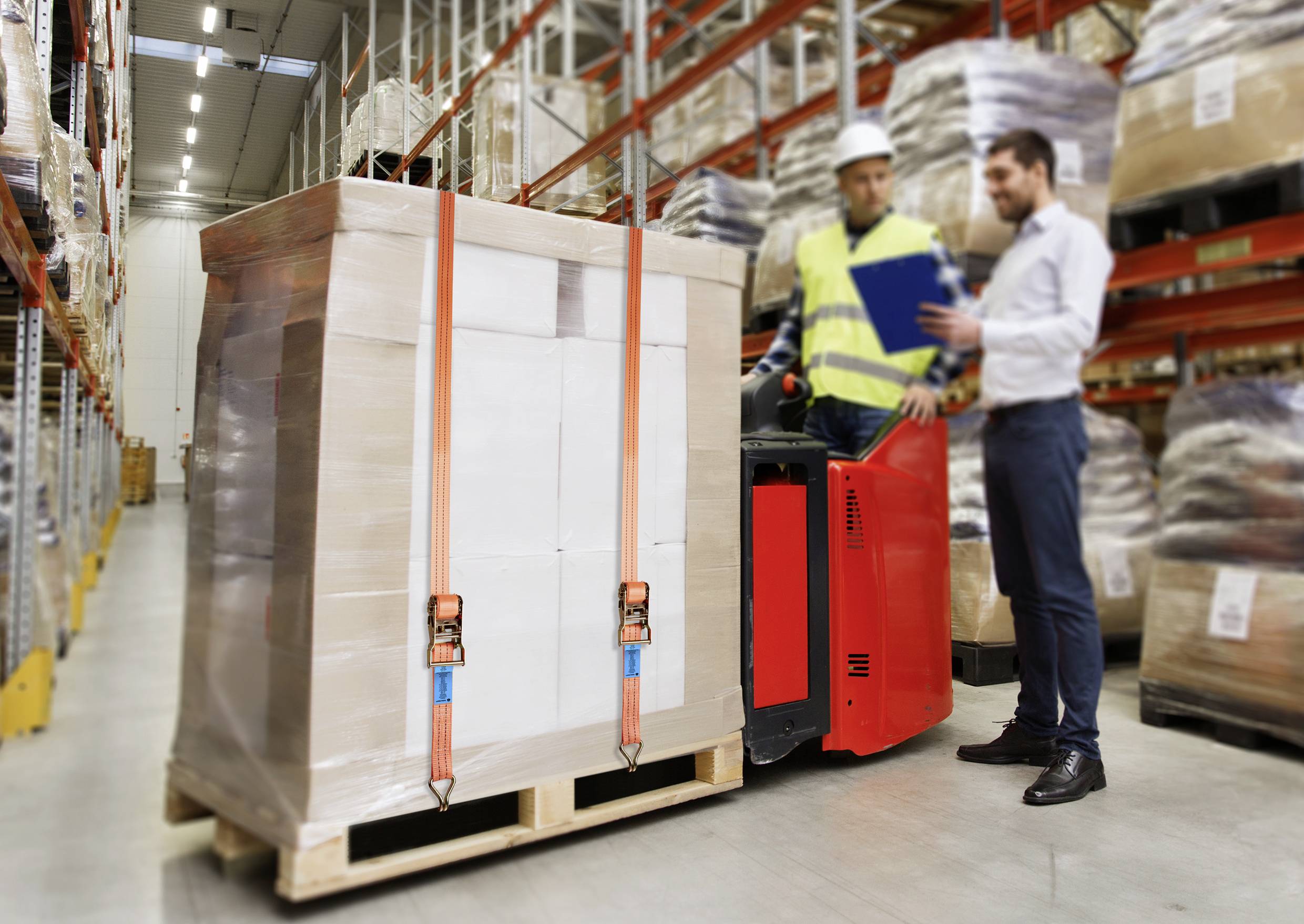 A pallet of cardboard boxes is being transported by a worker using a forklift in a warehouse. A manager is checking a clipboard.